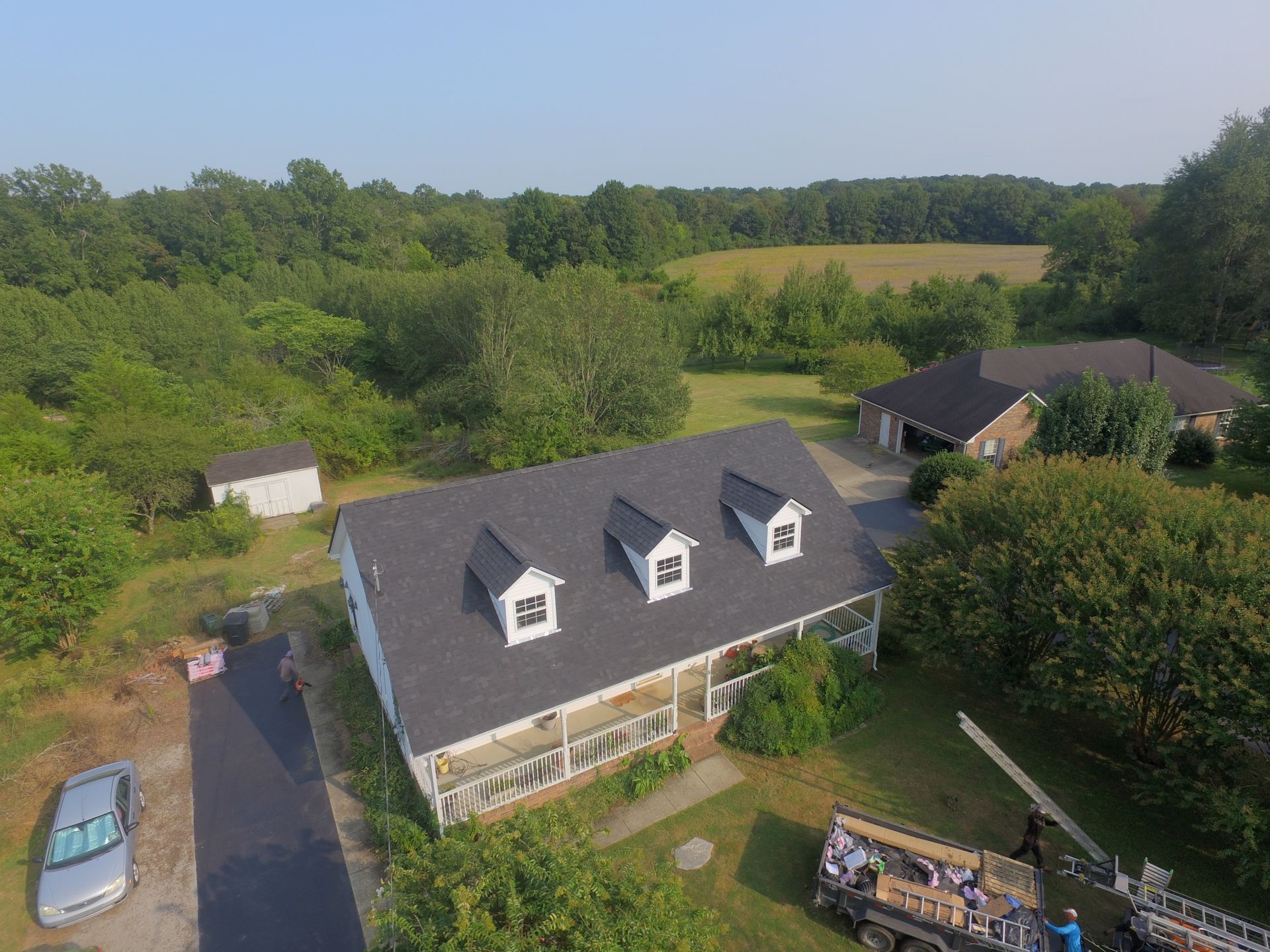 An aerial view of a white two-story house with a new dark gray roof and three dormers, surrounded by trees and a yard.