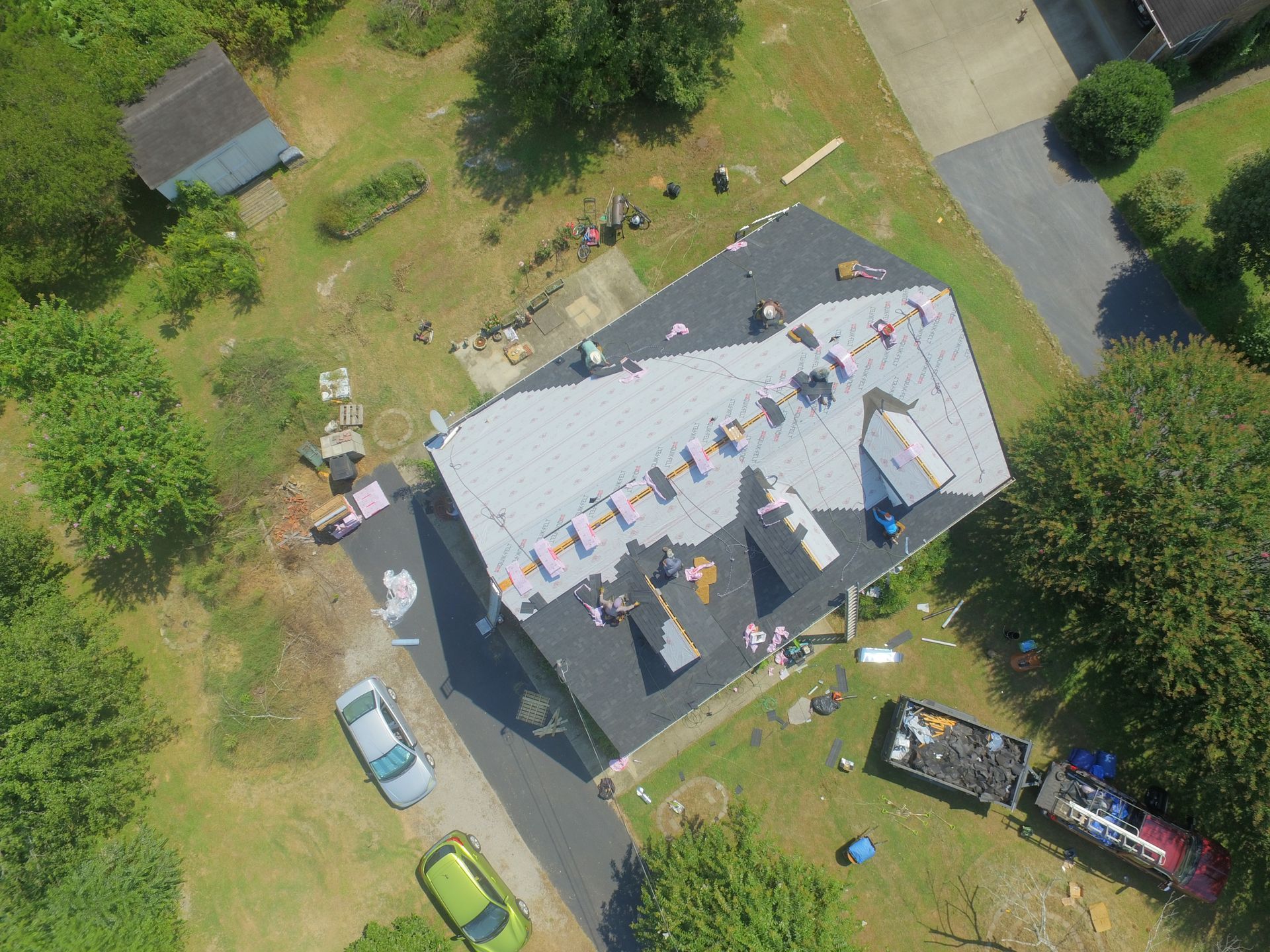 An aerial view of a house undergoing a roof replacement with workers, materials, and a debris trailer on the property.
