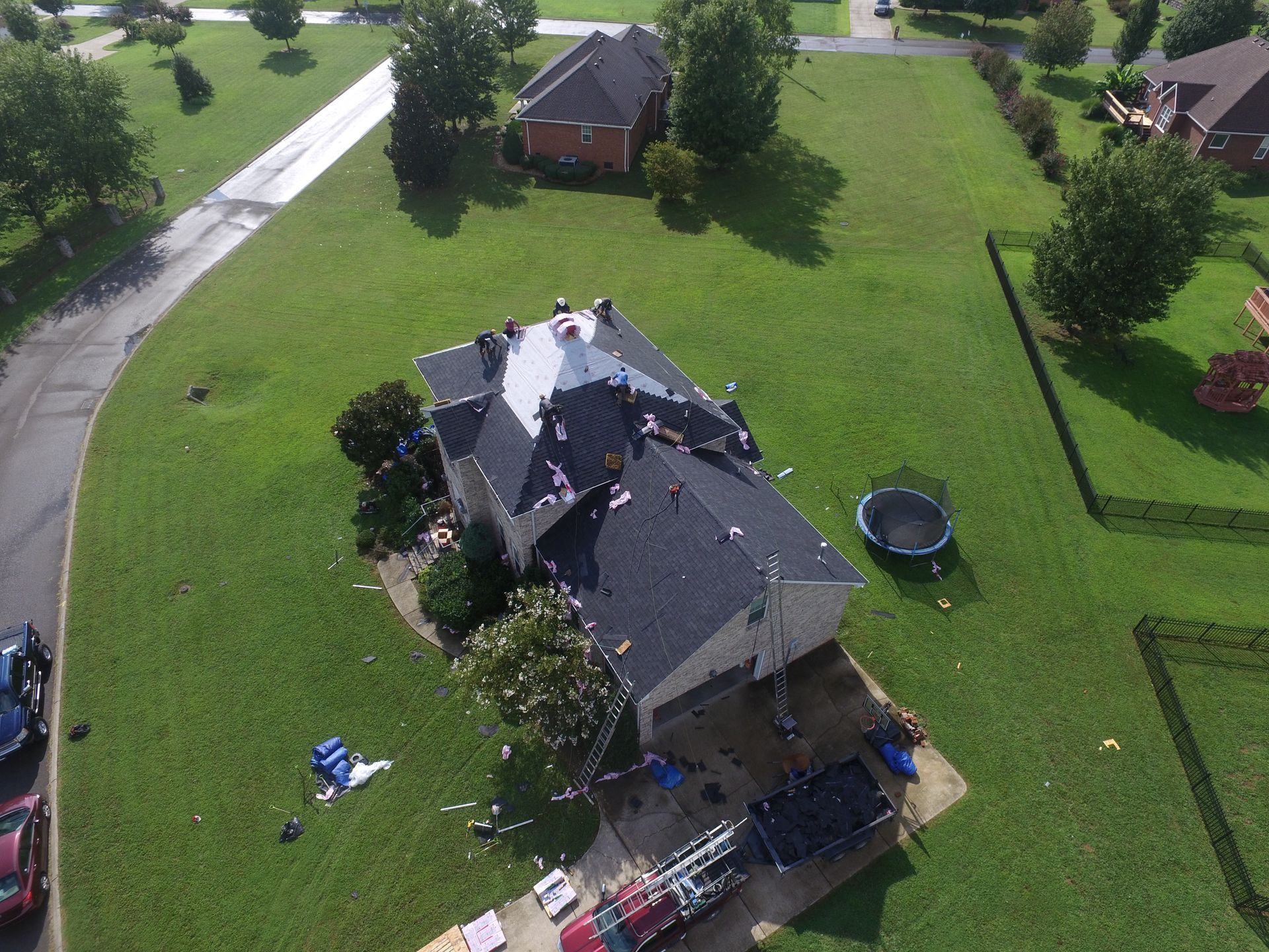 Aerial view of a residential house with a partially replaced black shingle roof, work equipment on the driveway and lawn.