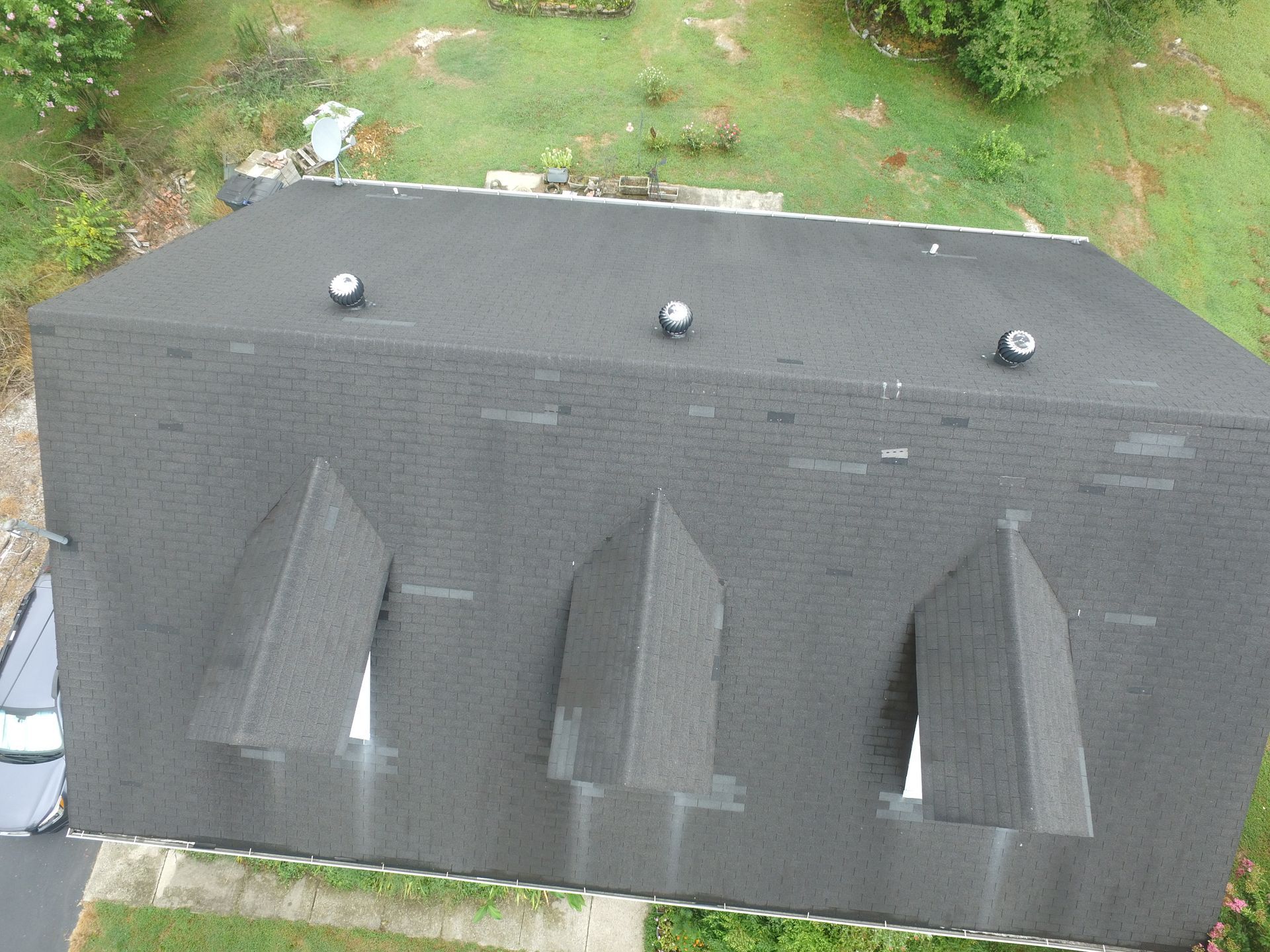 An aerial view of a dark shingled roof featuring three dormer windows and three roof vents.