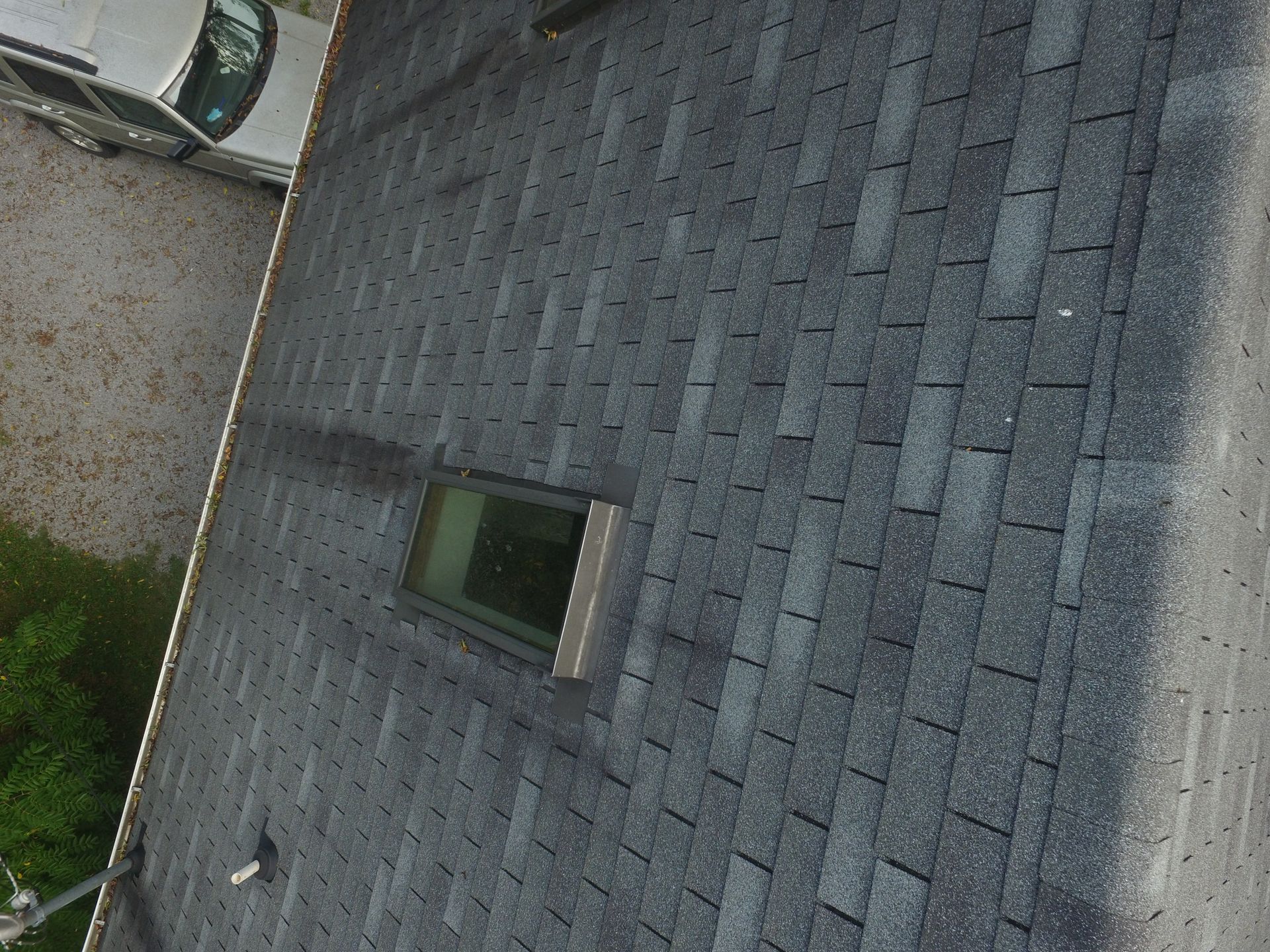 Aerial view of a gray asphalt shingle roof with a skylight, with part of a car visible on the gravel driveway below.