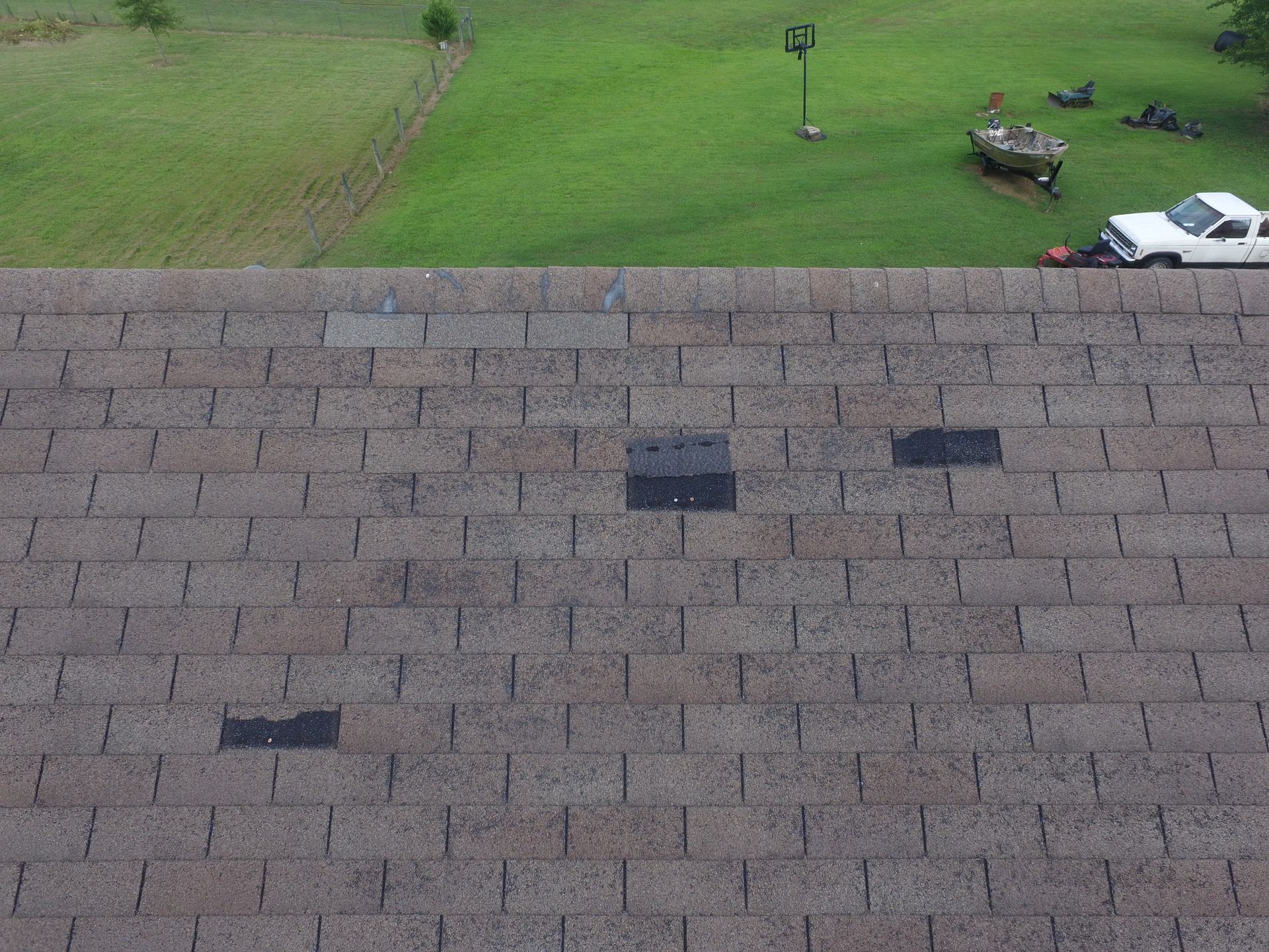 A high-angle view of a brown asphalt shingled roof with three black patches covering damaged or missing shingles.