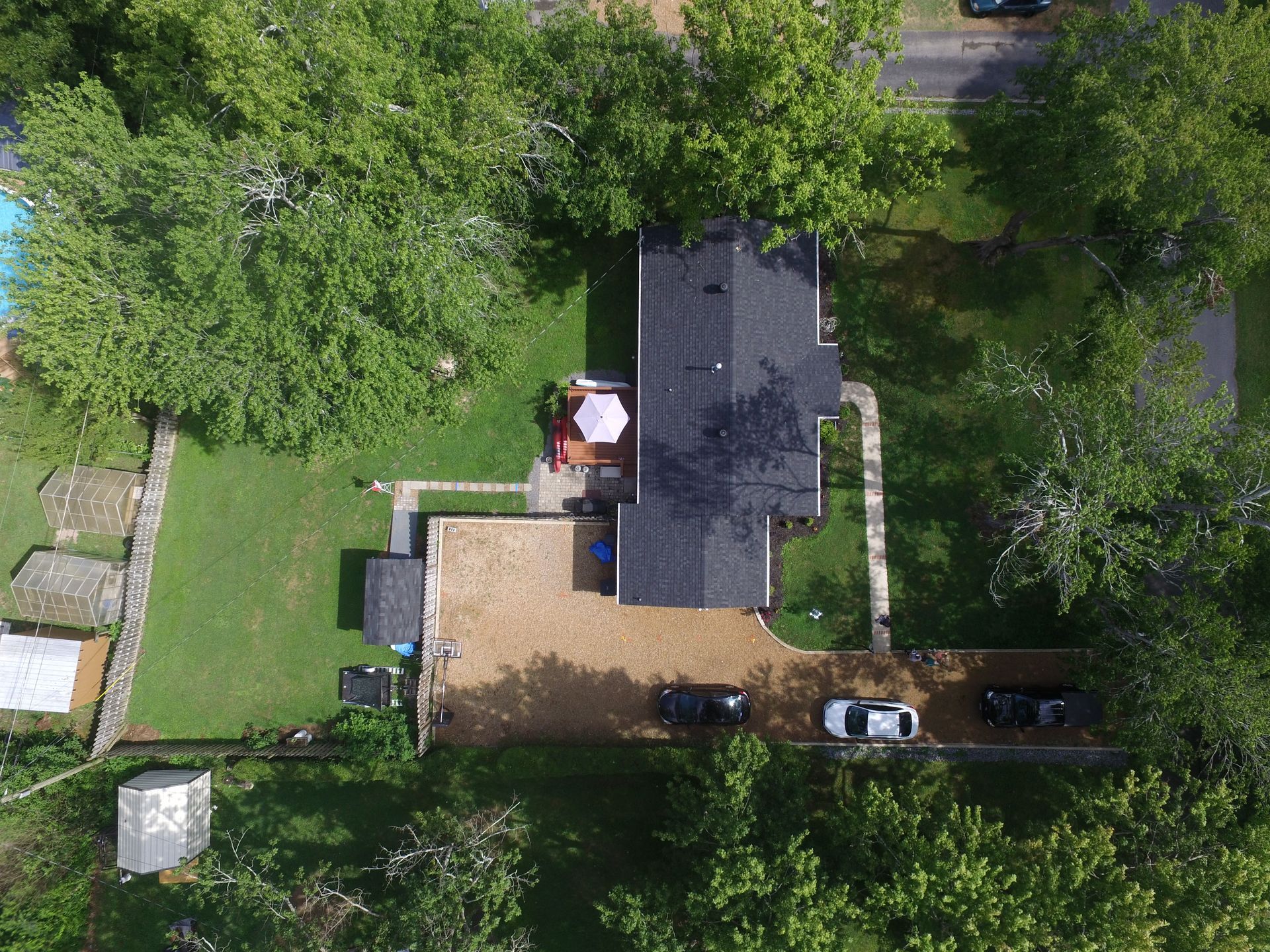 An aerial view of a house with a gravel driveway, three parked cars, a backyard with trees, and a patio with an umbrella.