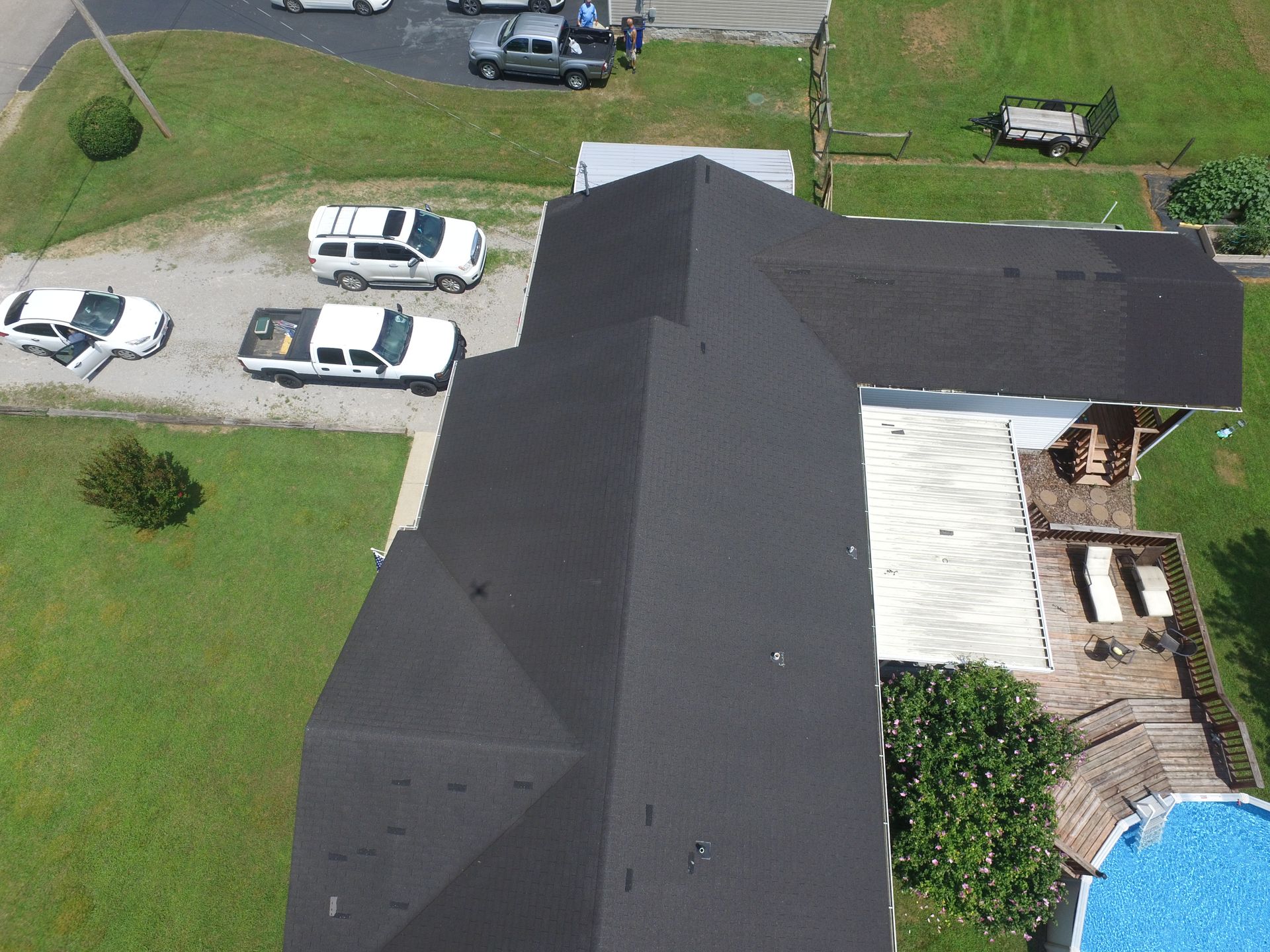 An aerial view of a dark-shingled roof on a house, with several vehicles parked on the grass and gravel driveway nearby.