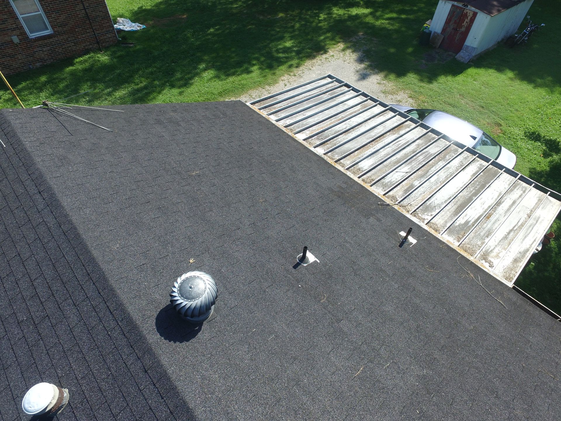 An aerial view of a dark shingled roof with a vent, plumbing stacks, and a partially uncovered, weathered wooden section.