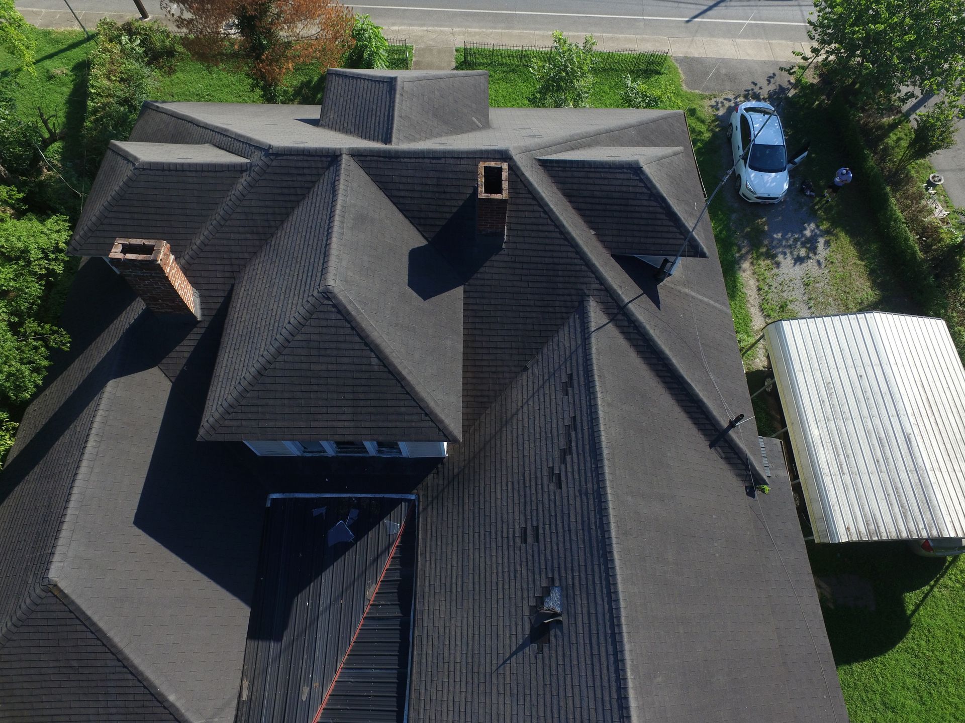 Aerial view of a complex, multi-gabled dark shingled roof with two chimneys, next to a driveway and small white structure.