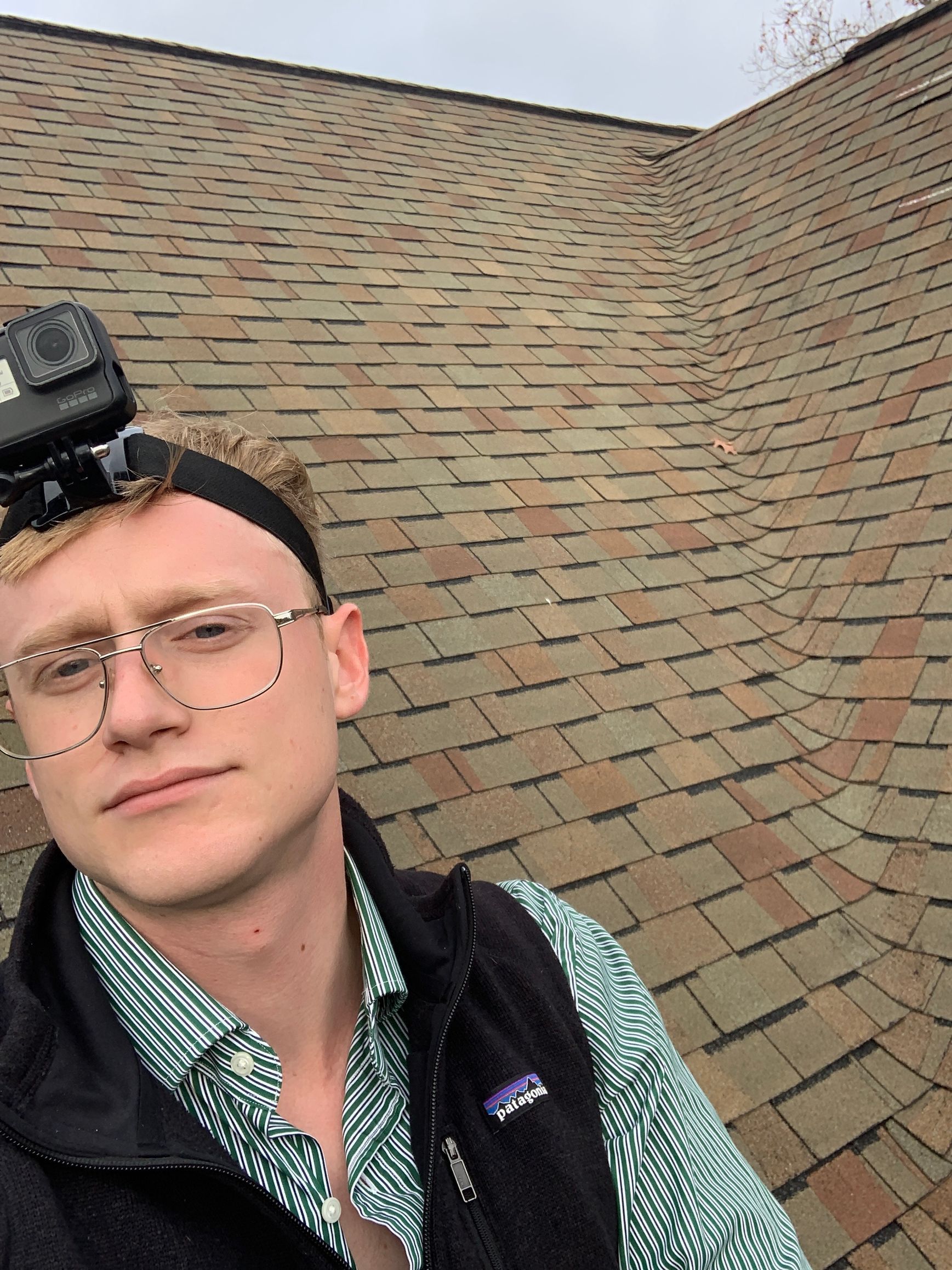A person wearing a head-mounted camera and glasses stands in front of a shingled roof.
