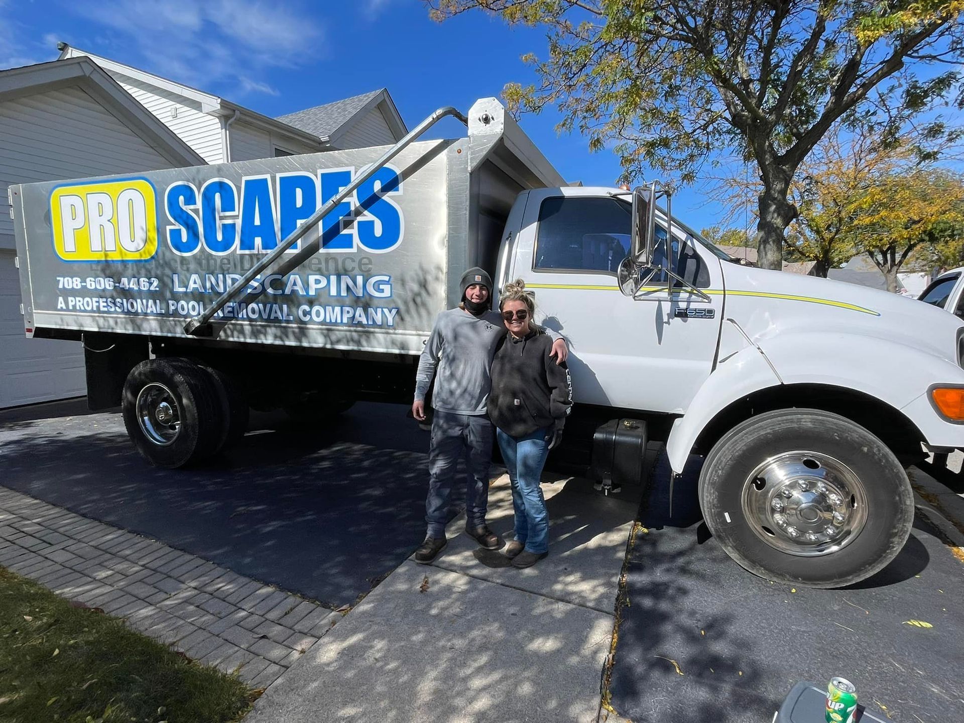 Two people stand in front of a white Pro Scapes dump truck parked on a paved driveway near a house.