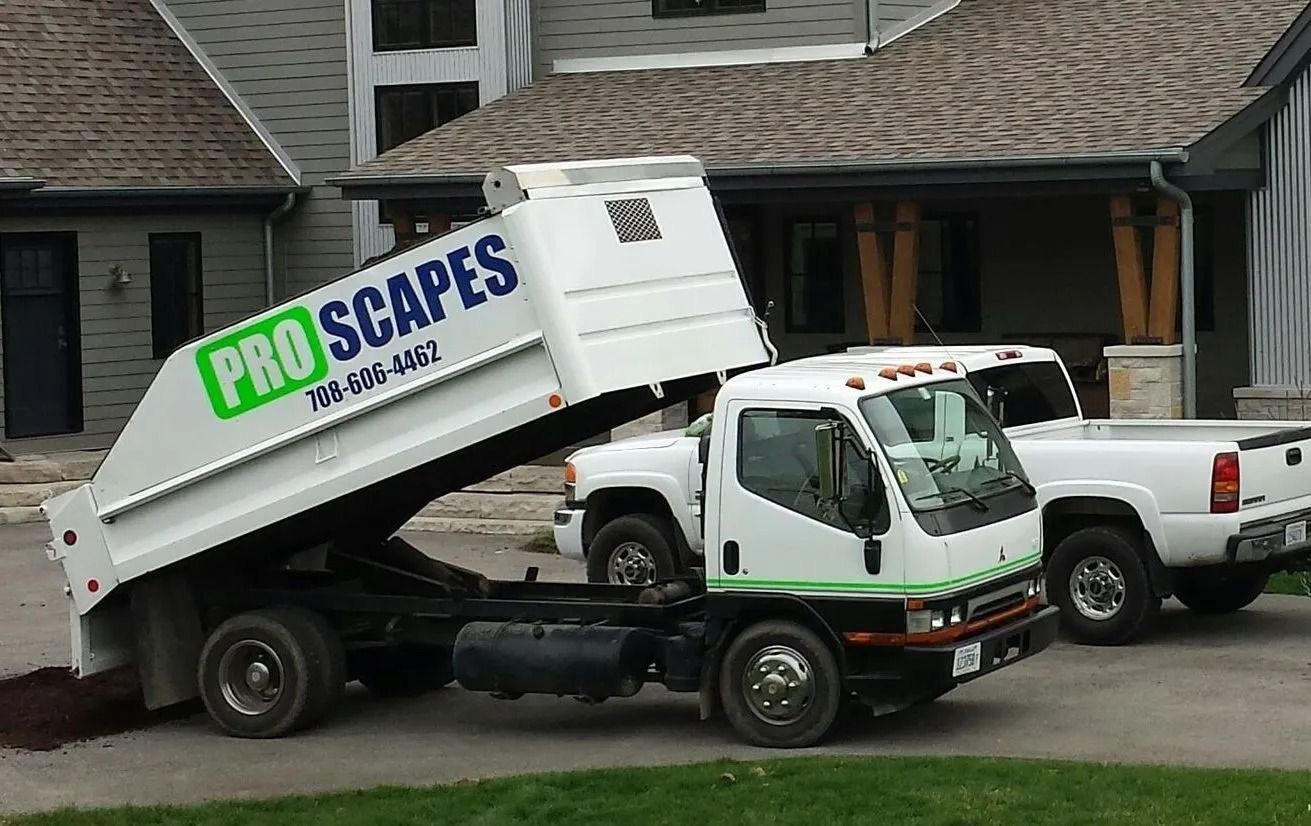 A white ProScapes dump truck with its bed raised, parked in a driveway in front of a house.