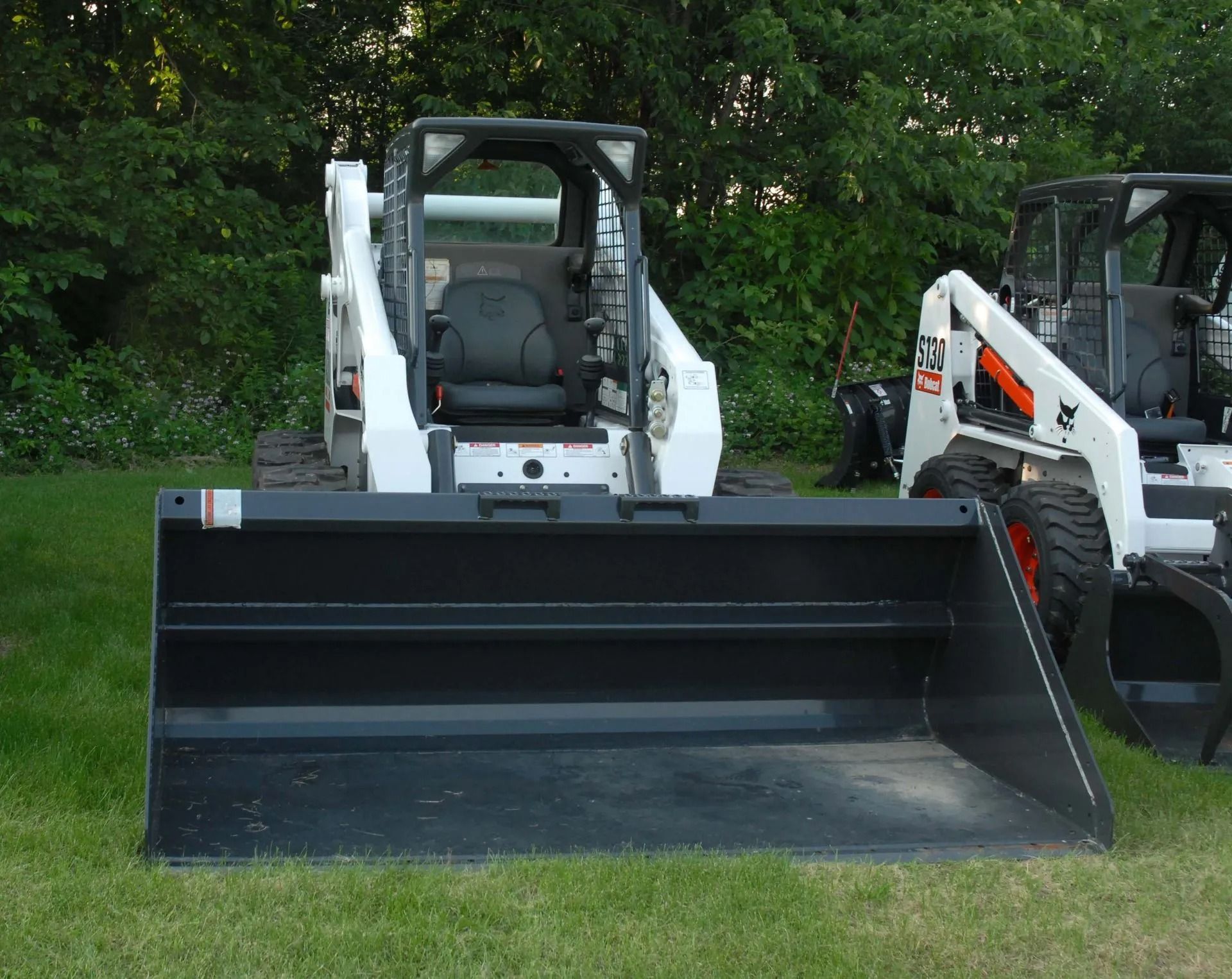 A large black bucket attachment sits on the grass in front of two white Bobcat skid-steer loaders parked outdoors.