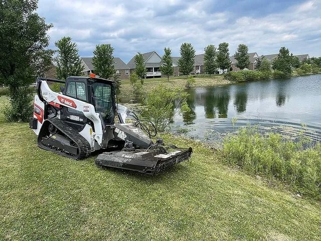 A white and black Bobcat compact track loader with a brush cutter mower attachment parked near a suburban pond.