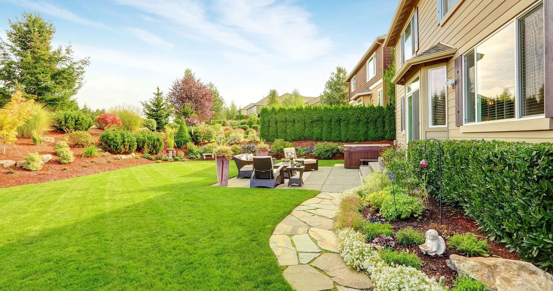 A backyard with a stone path, a grassy lawn, patio furniture, and a landscaped garden next to a beige house.
