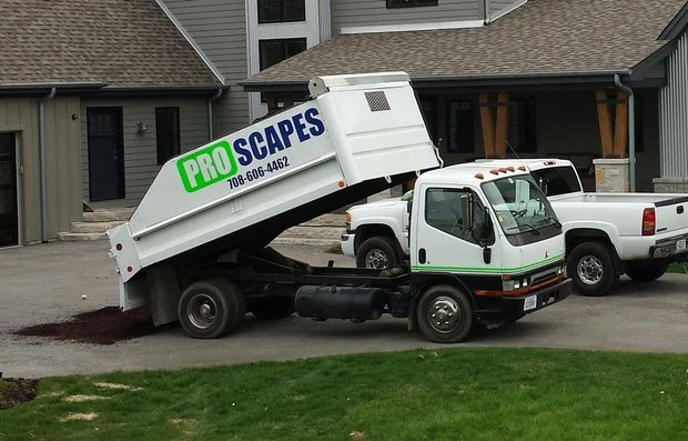A white Proscapes dump truck parked in a driveway, elevated to unload soil in front of a house.