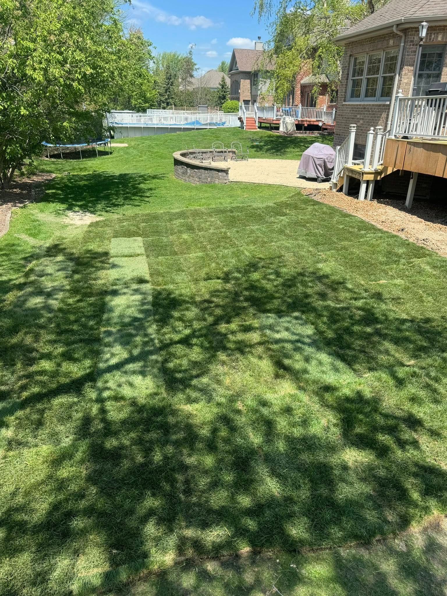 A residential backyard featuring newly laid sod patches on a grassy lawn near a stone retaining wall and wooden deck.