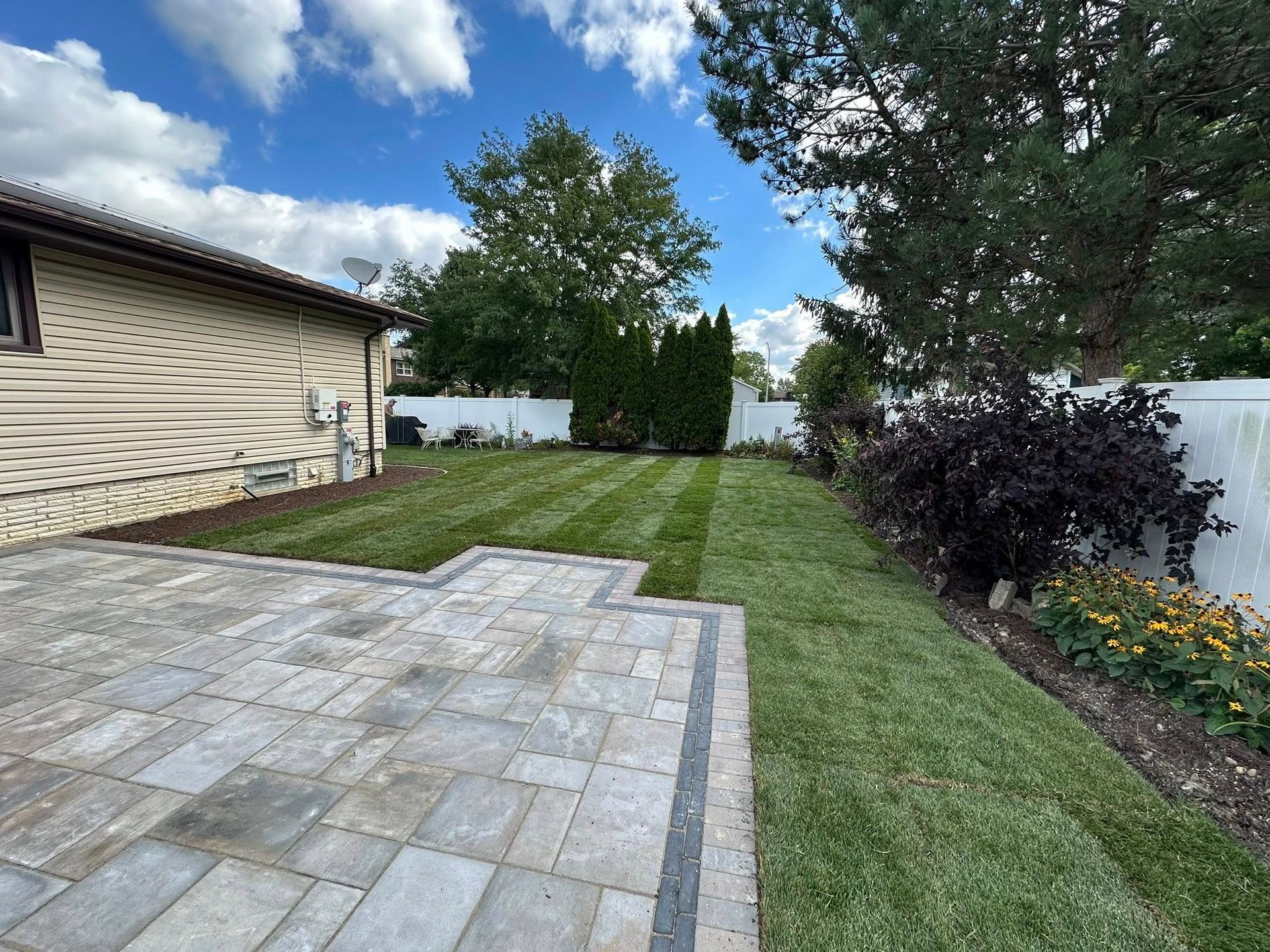 A paved stone patio leads to a manicured green lawn in a suburban backyard, bordered by a house and lush landscaping.