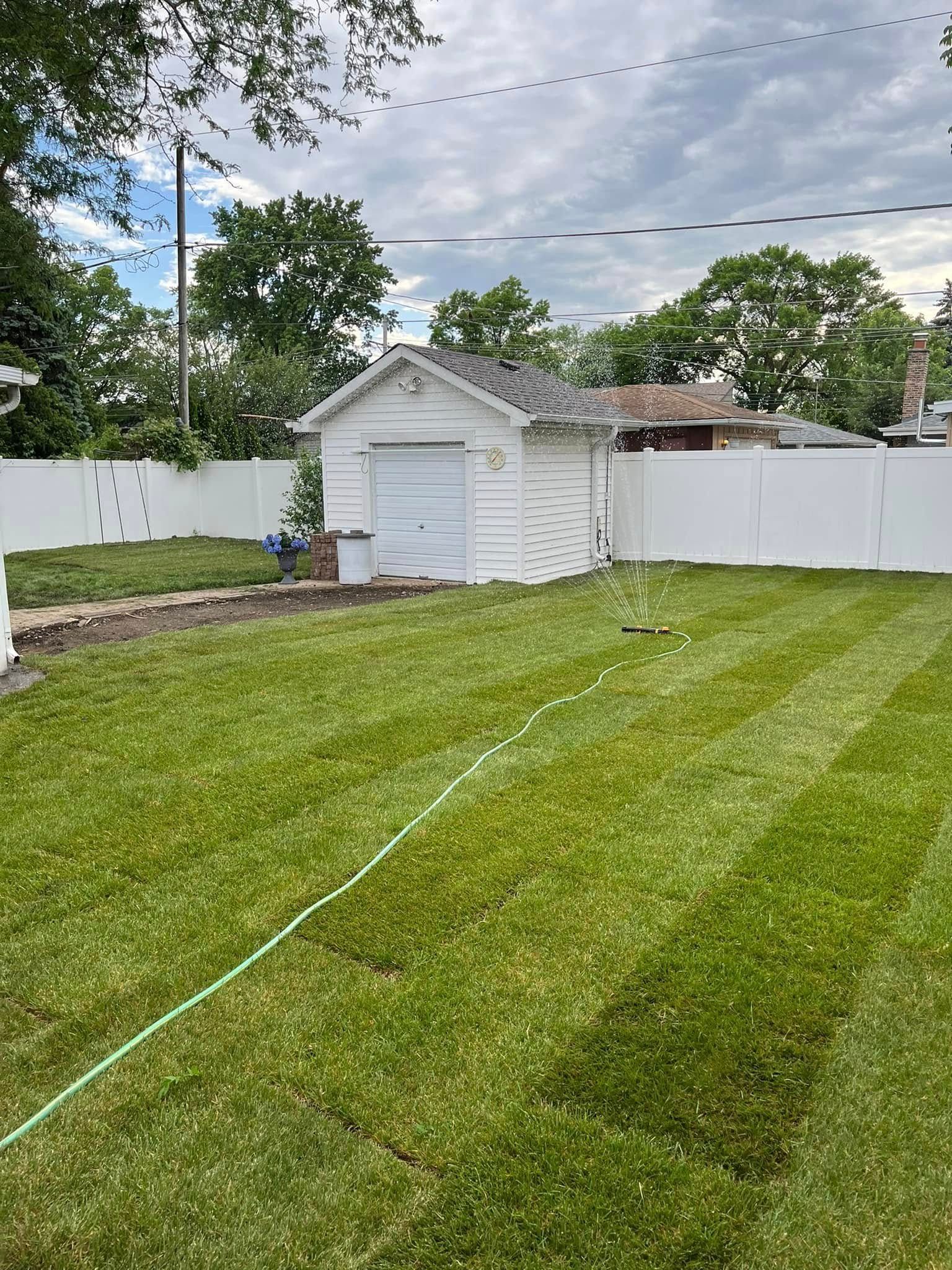 A bright green backyard with a white detached garage, a white vinyl fence, and a hose spraying water on the lawn.