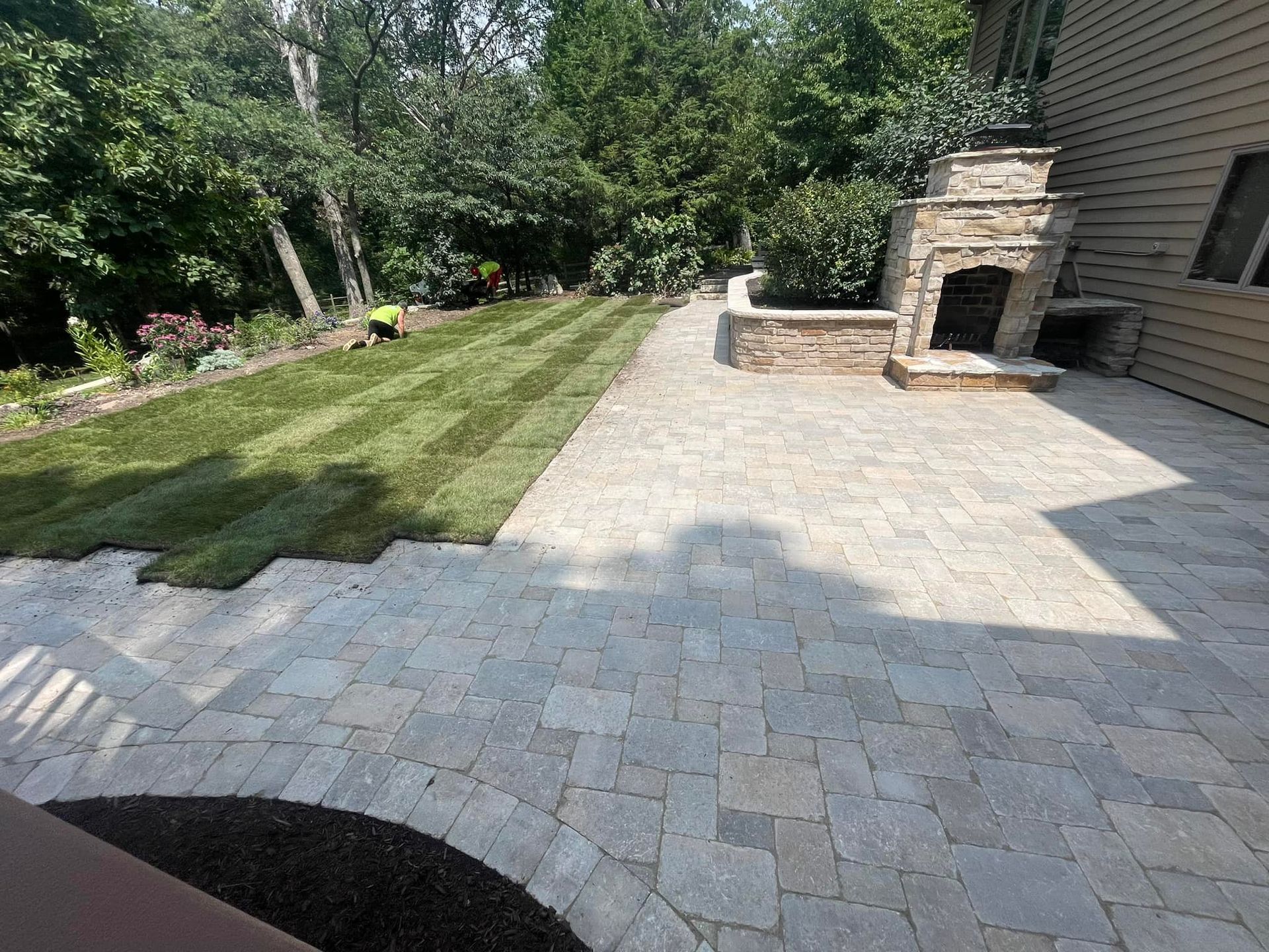 A patio paved with gray stones borders a freshly laid green lawn next to a stone outdoor fireplace and a wooden house.