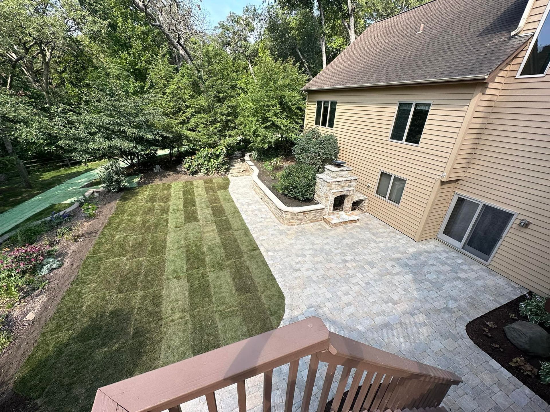 High-angle view of a tan house with a large stone patio, a small outdoor fireplace, and a freshly mowed green lawn.