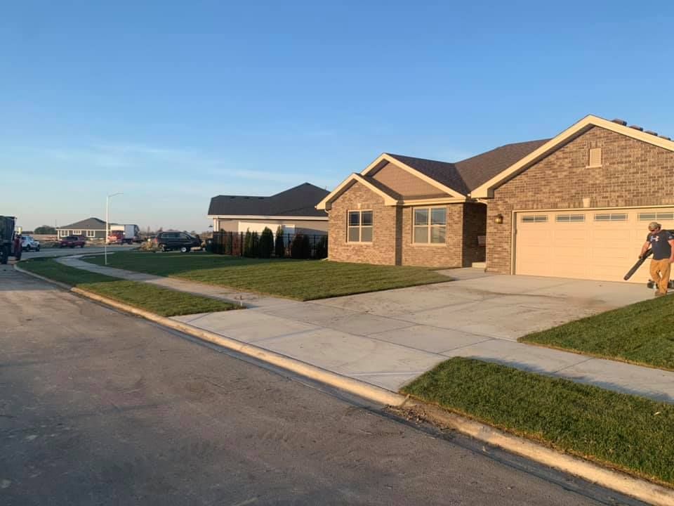A person uses a leaf blower on the driveway of a tan brick suburban house on a sunny day.