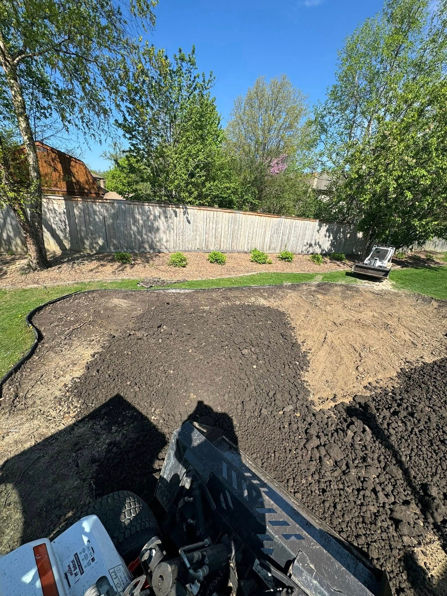 A view from a skid steer loader shows a freshly leveled patch of dark, rich soil in a sunny backyard with a fence and trees.