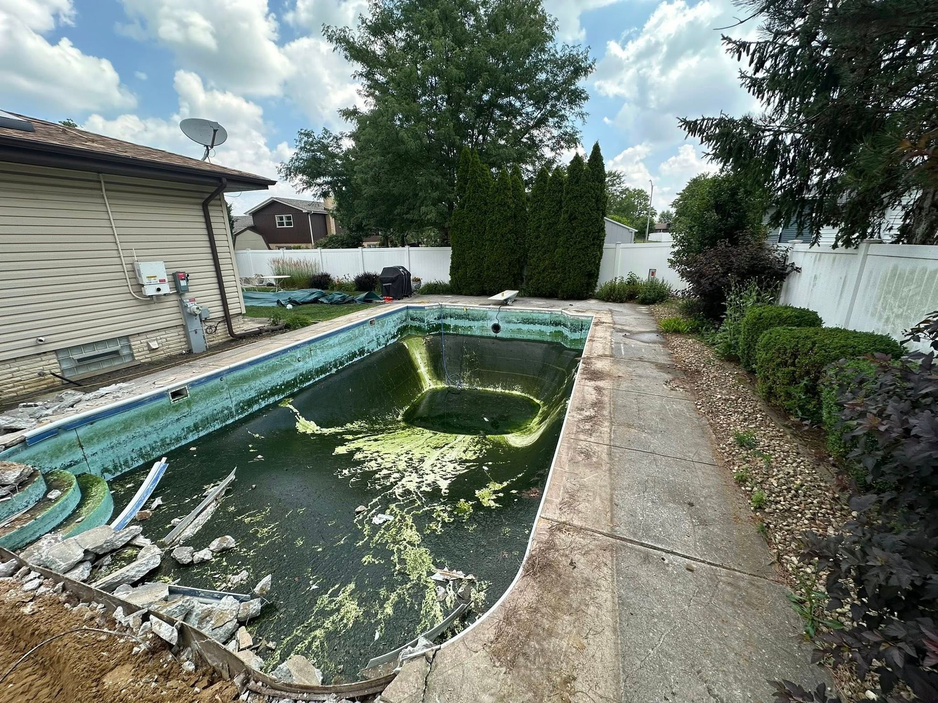 A neglected in-ground swimming pool with murky water and debris, located in a backyard next to a residential house.