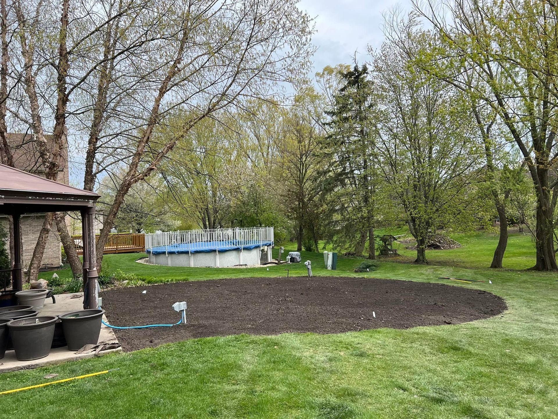 A circular patch of dark dirt in a grassy backyard, with a gazebo on the left and an above-ground pool in the distance.
