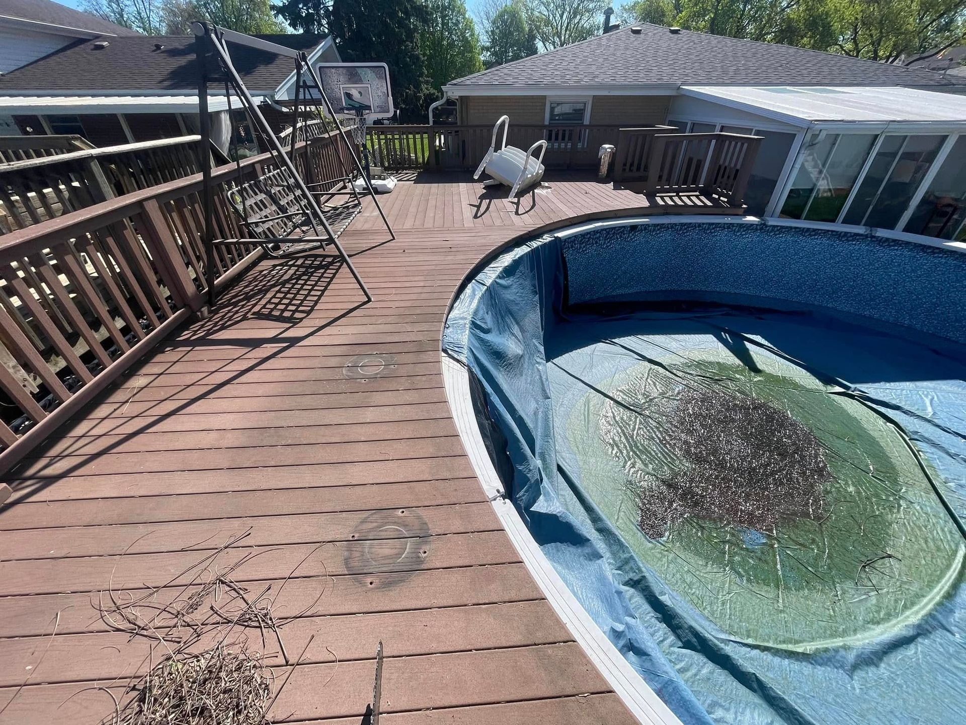 A wooden deck surrounds an above-ground pool filled with cloudy, stagnant water and debris on a sunny day.