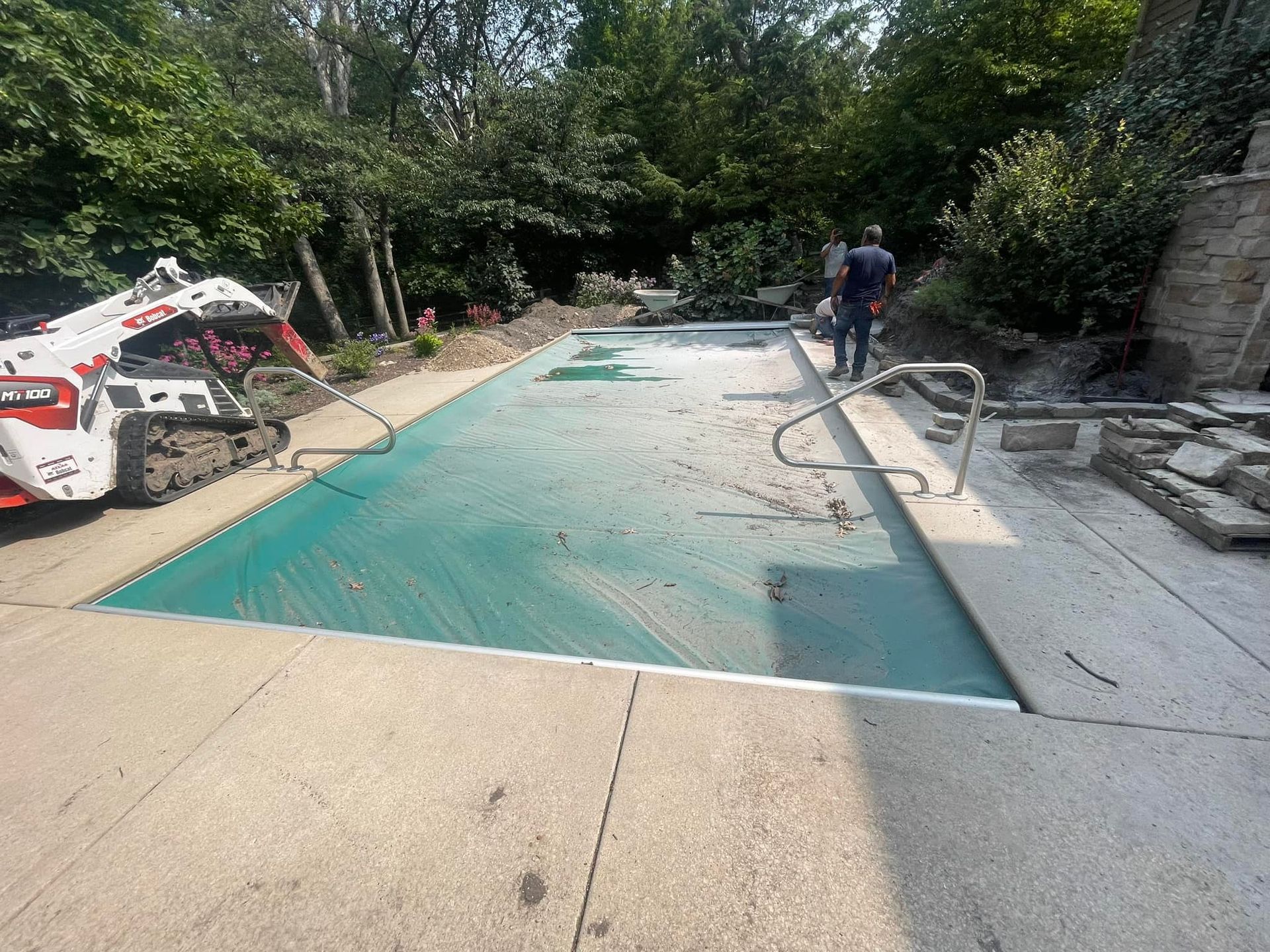 A skid steer loader parked next to a rectangular swimming pool covered with a green tarp in a backyard setting.