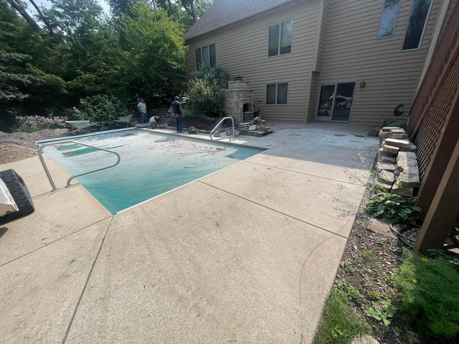 A backyard swimming pool with a safety cover sits beside a multi-story house and a concrete patio with construction work.
