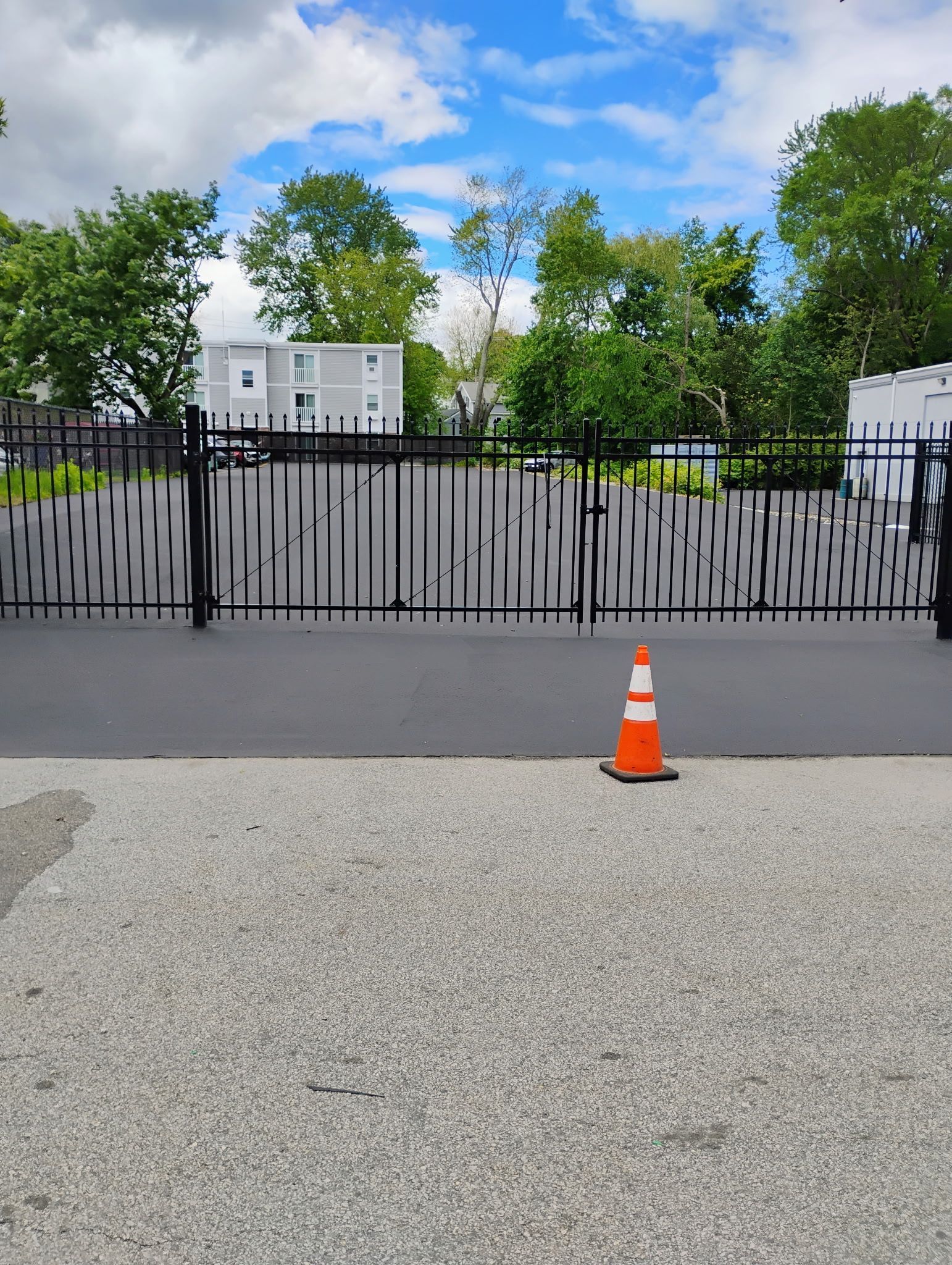 Black metal gate blocking a roadway, with an orange traffic cone in front and buildings behind trees.