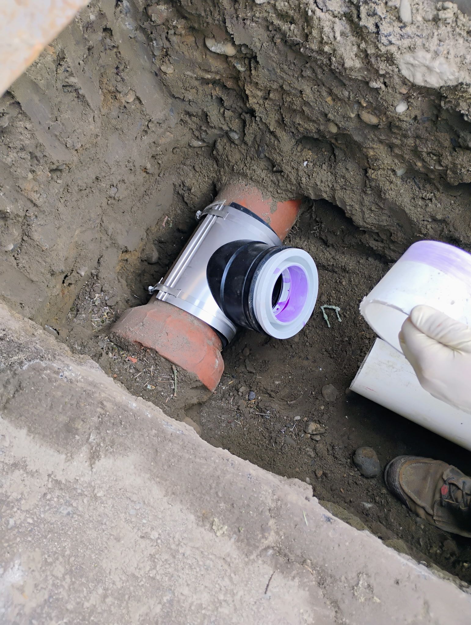 Underground plumbing pipe connection with white and purple fittings in a dirt trench