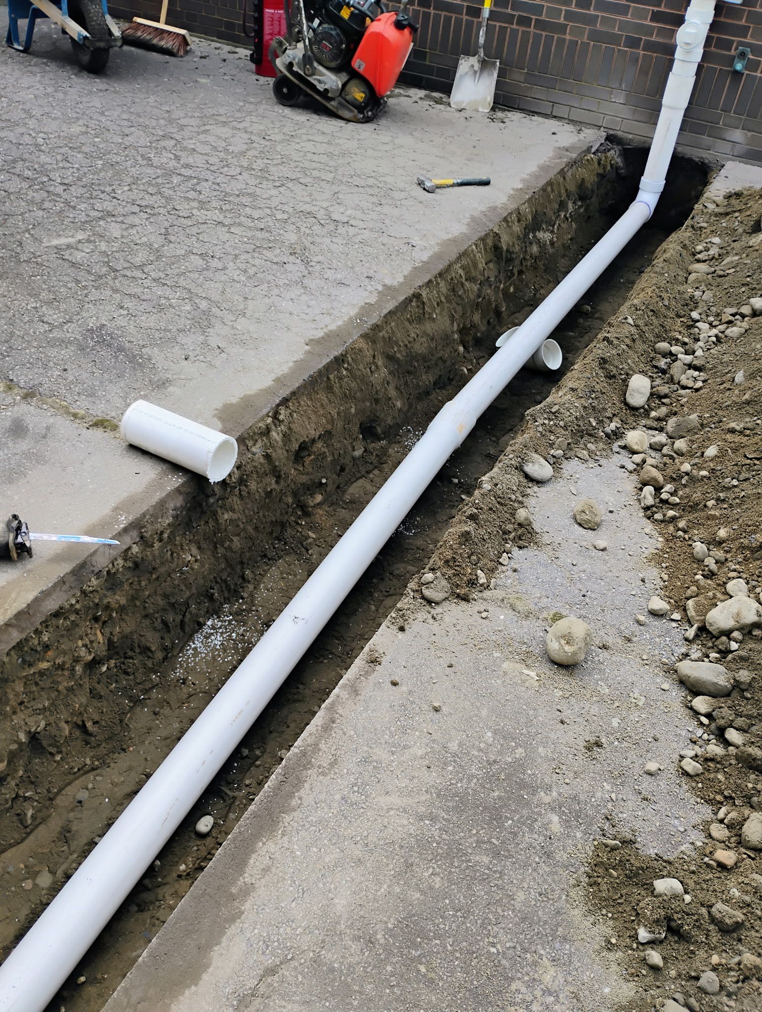 White pipe laid in a roadside trench beside a gravel path and concrete curb, with construction equipment in the background