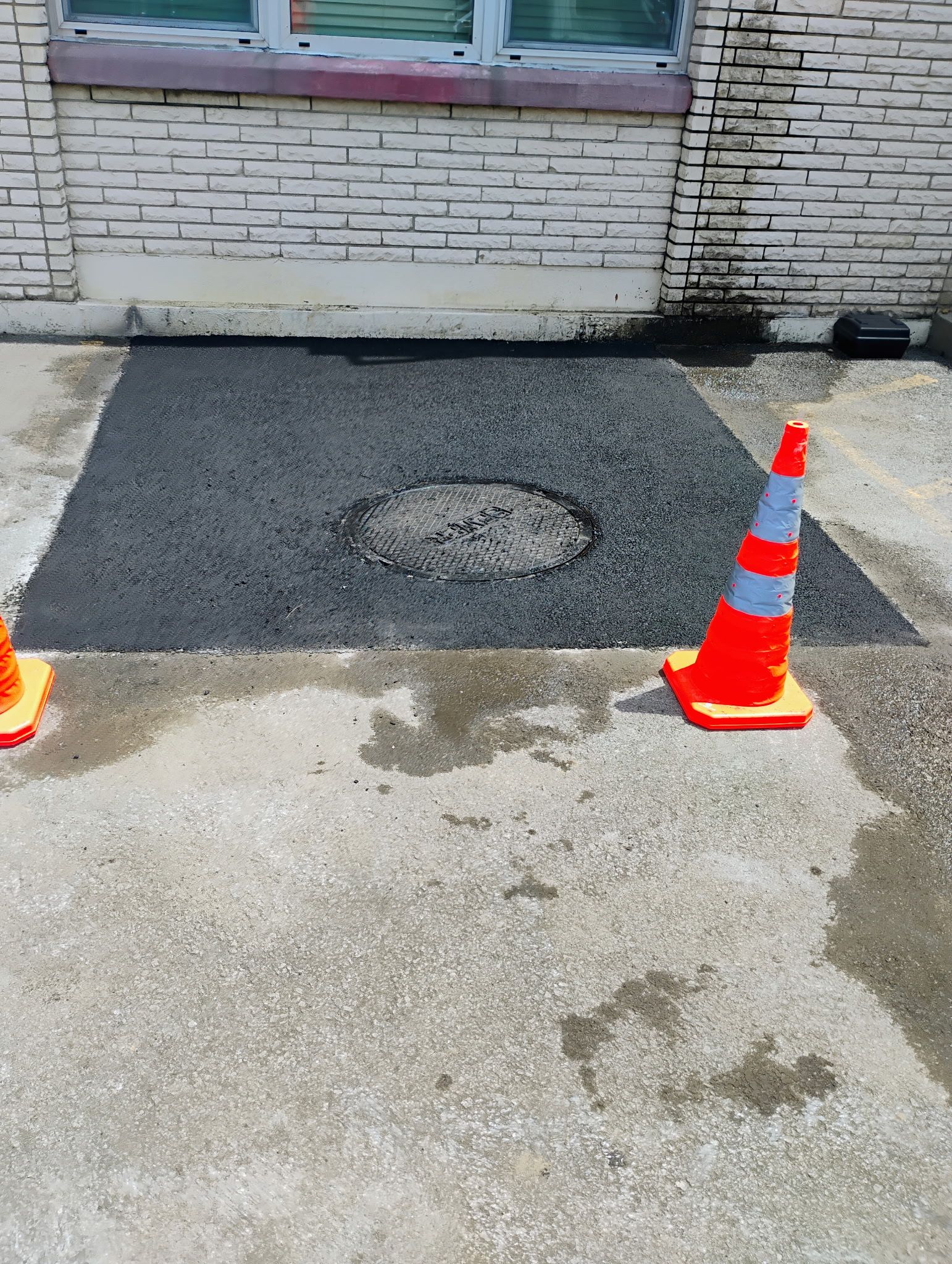 Orange traffic cones flank a rough asphalt patch beside a brick building wall.