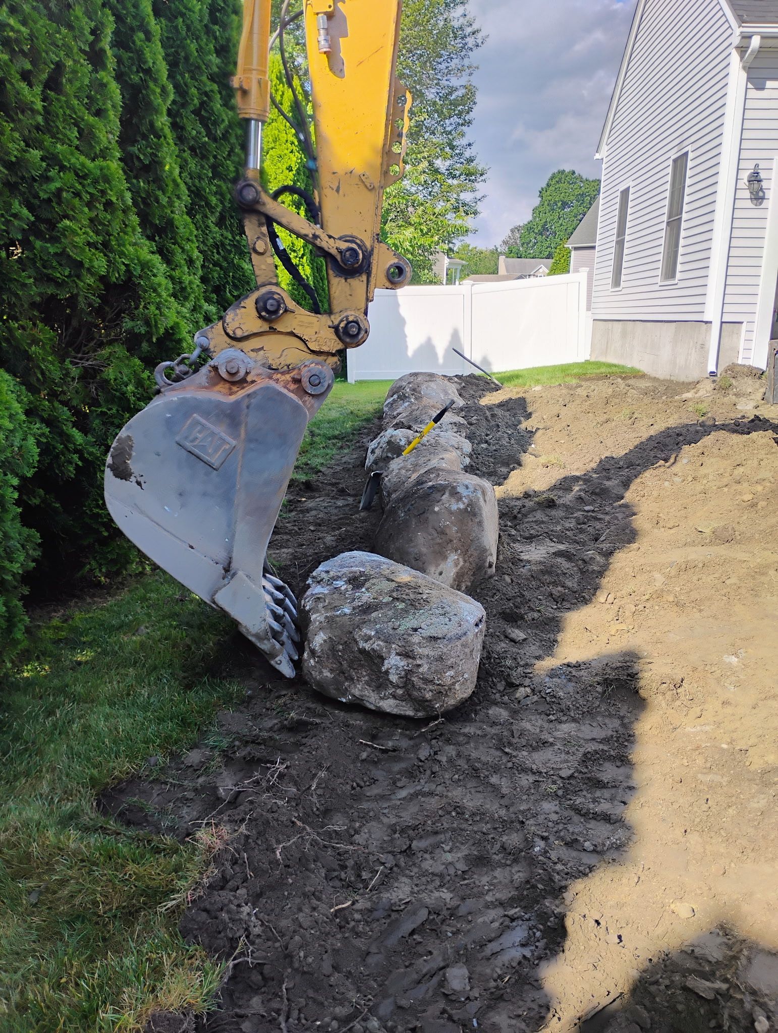 Excavator clearing dirt beside a large rock in a landscaped yard near a white house.