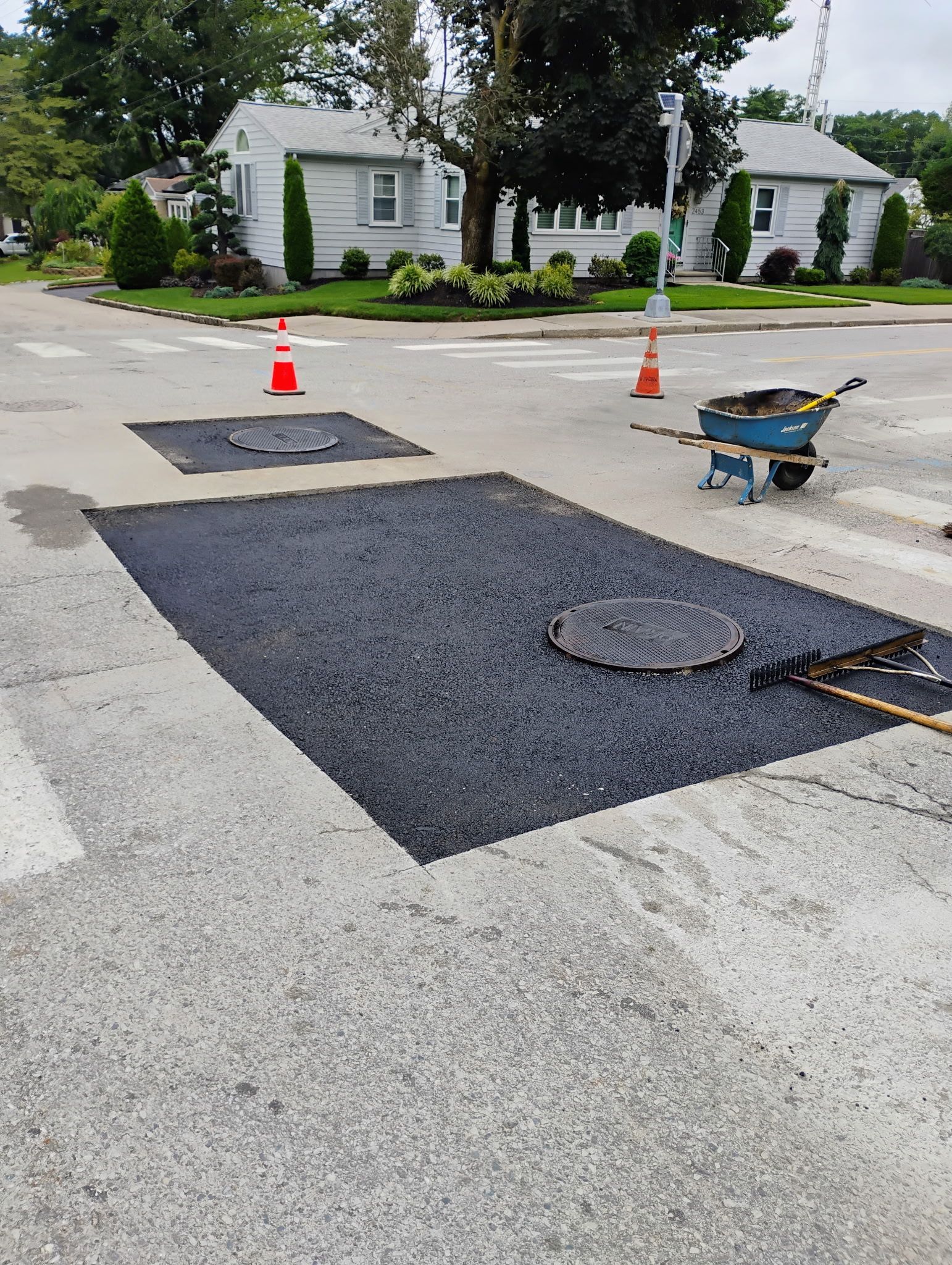 Roadwork patch on residential street with asphalt squares, orange cones, and a wheelbarrow.