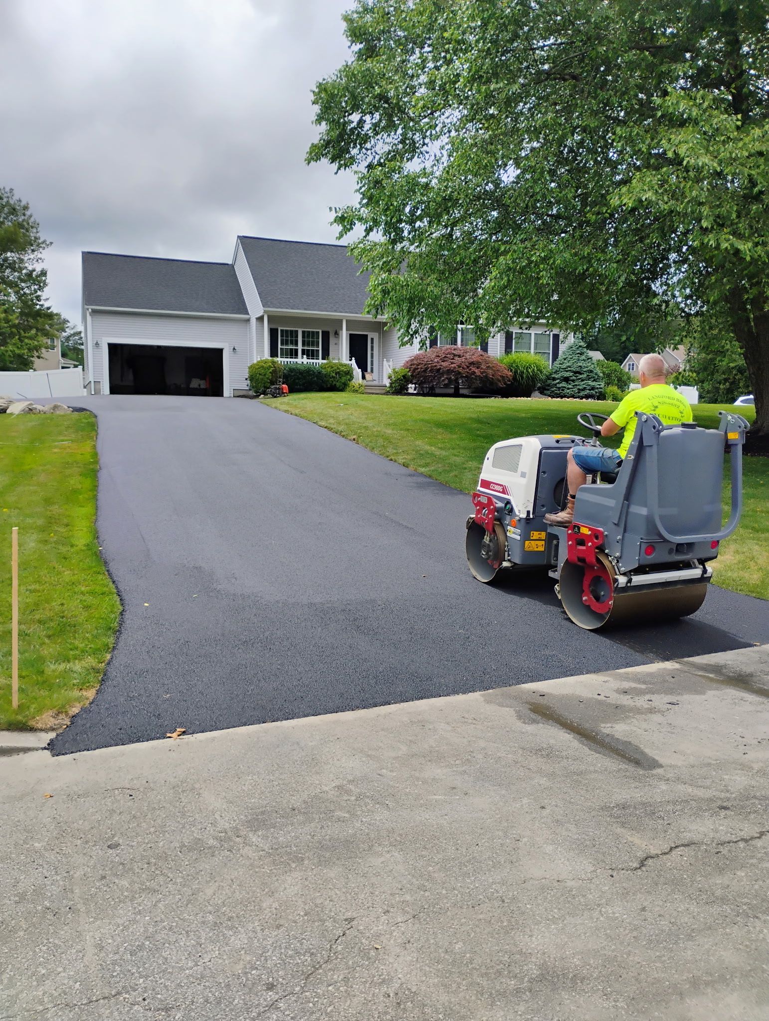 Worker operating a ride-on lawn mower on a freshly paved driveway in front of a suburban house.