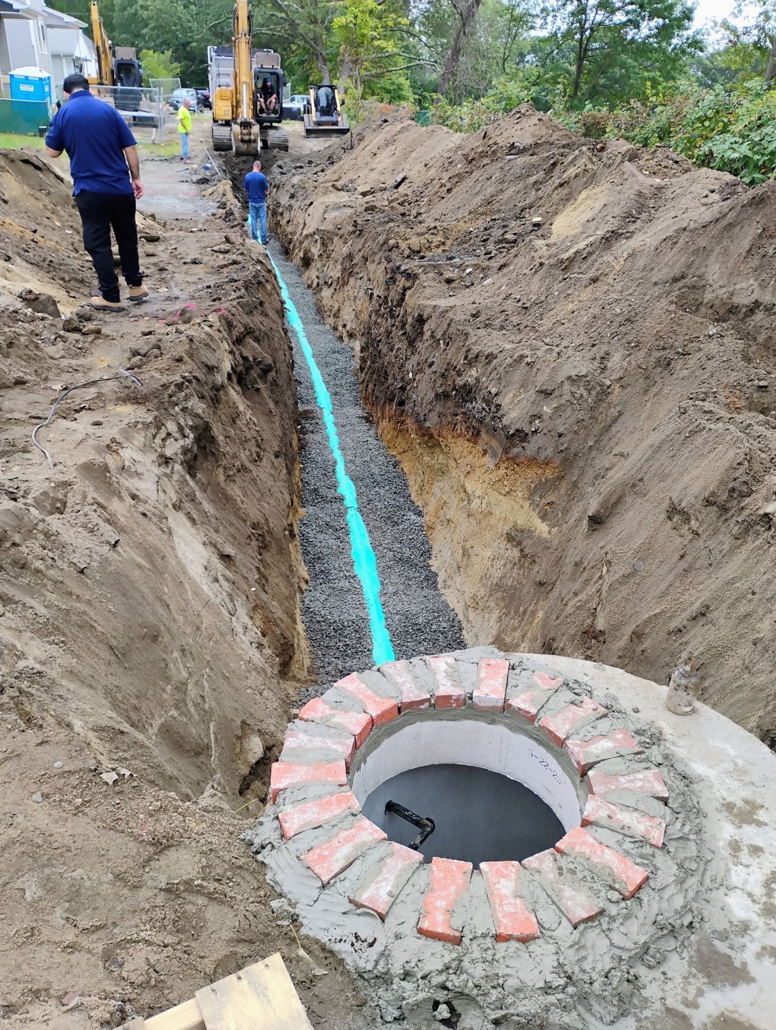 Construction trench with blue pipe laid in gravel, concrete manhole ring, and workers in the background