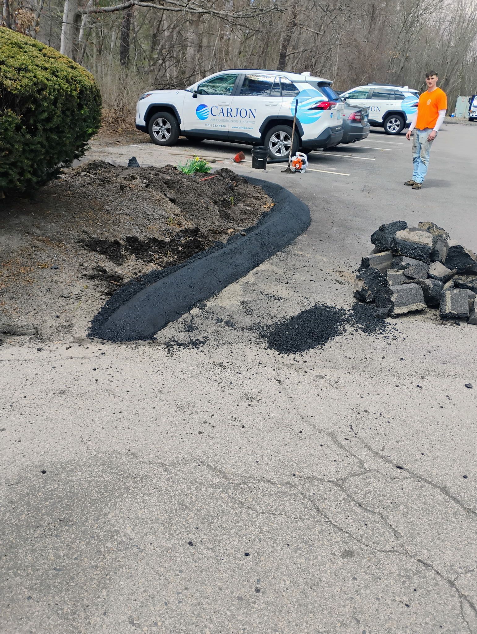 Roadside construction area with an SUV, worker in orange jacket, fresh black asphalt patch, and piled rocks