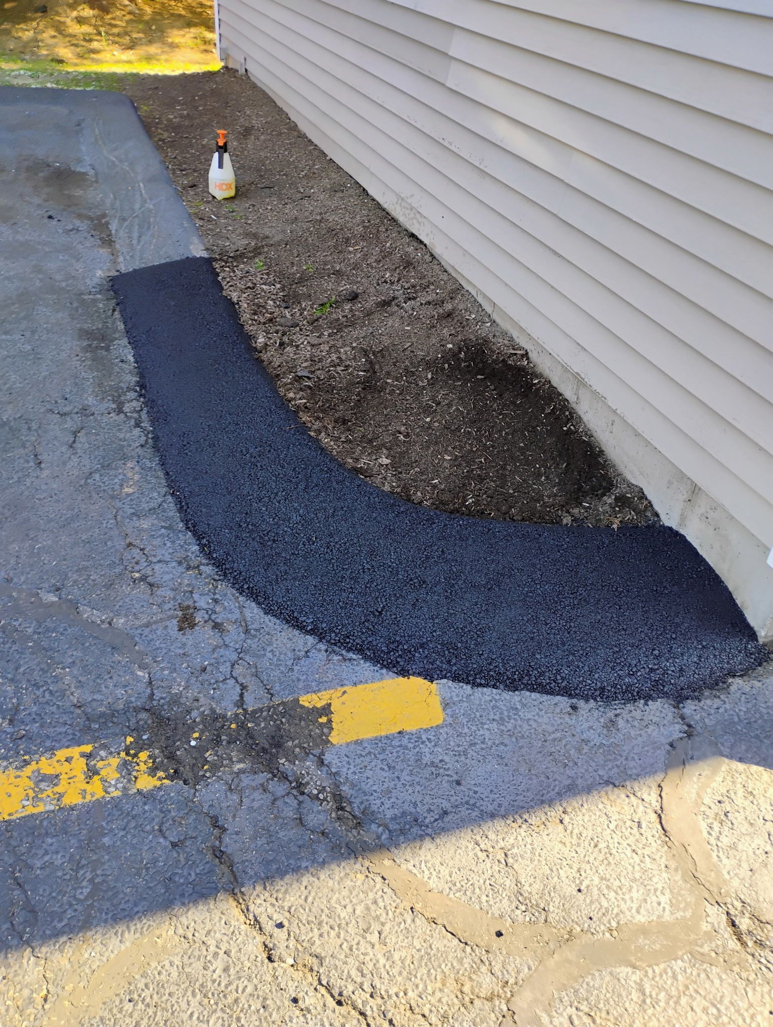 Cracked asphalt patch beside a building, with yellow curb markings and a small bottle near the wall