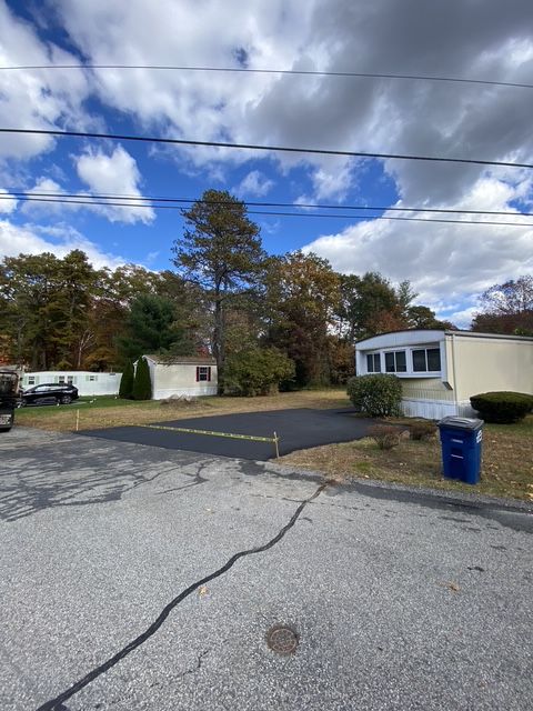 Rural street with mobile homes, cracked pavement, blue trash bin, and trees under a cloudy sky.