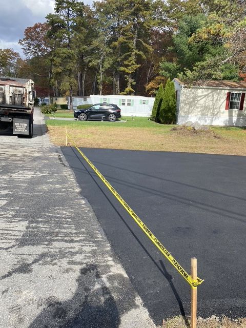 Fresh black asphalt driveway beside a gravel lot, with yellow caution tape and a parked car near a white trailer.