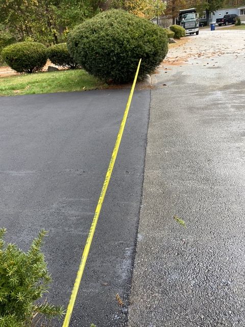 Yellow tape marks the edge between a paved driveway and asphalt near a large bush.