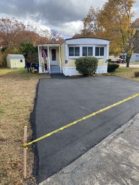Small white house with a front porch and a freshly paved black driveway in a suburban yard.