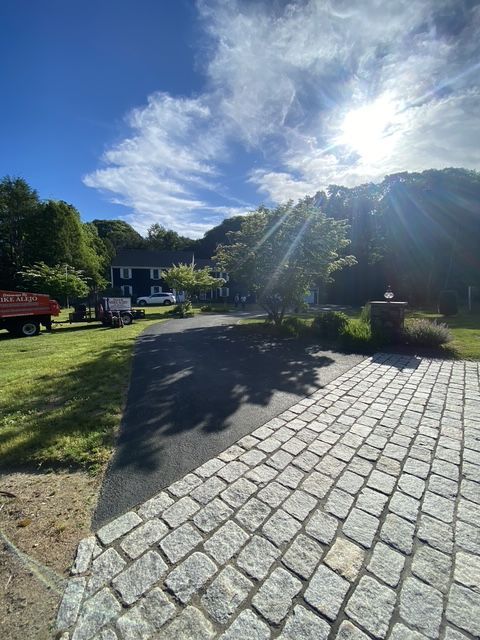 Sunlit cobblestone driveway beside a house and green lawn, with trees and blue sky in the background