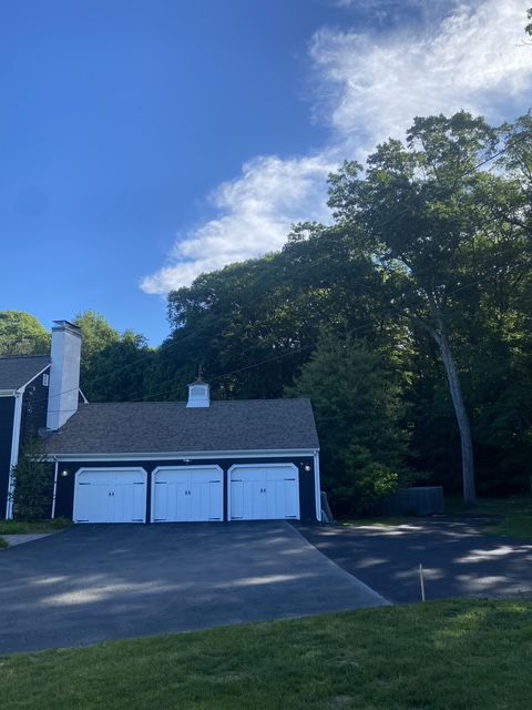 White garage with three doors beside a driveway and tall trees under a blue sky.