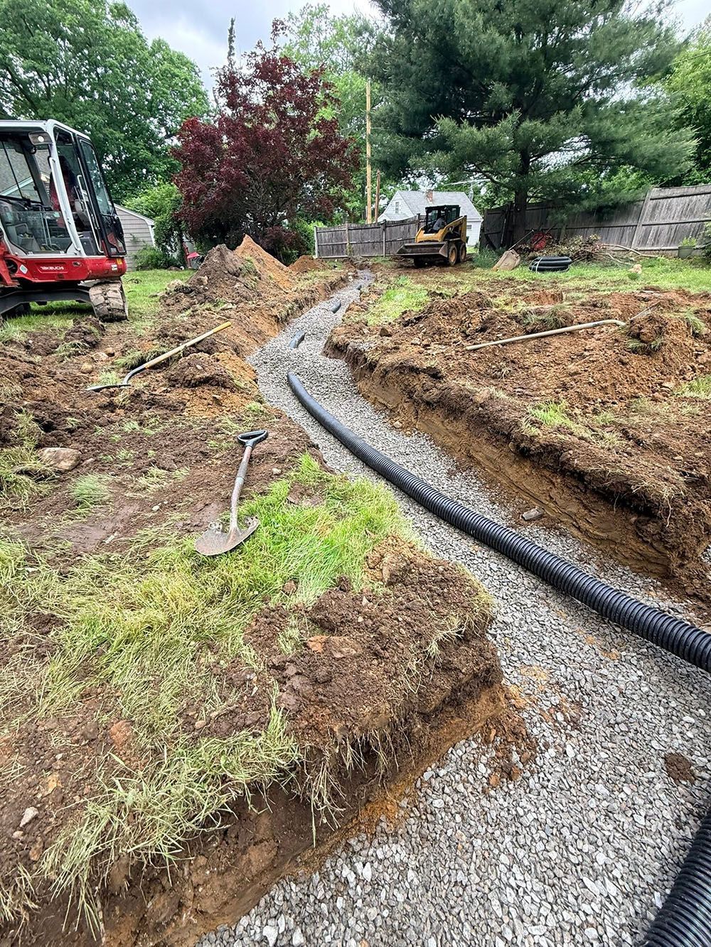 Construction site with gravel trench, black pipe, excavator, and dirt piles under overcast sky.
