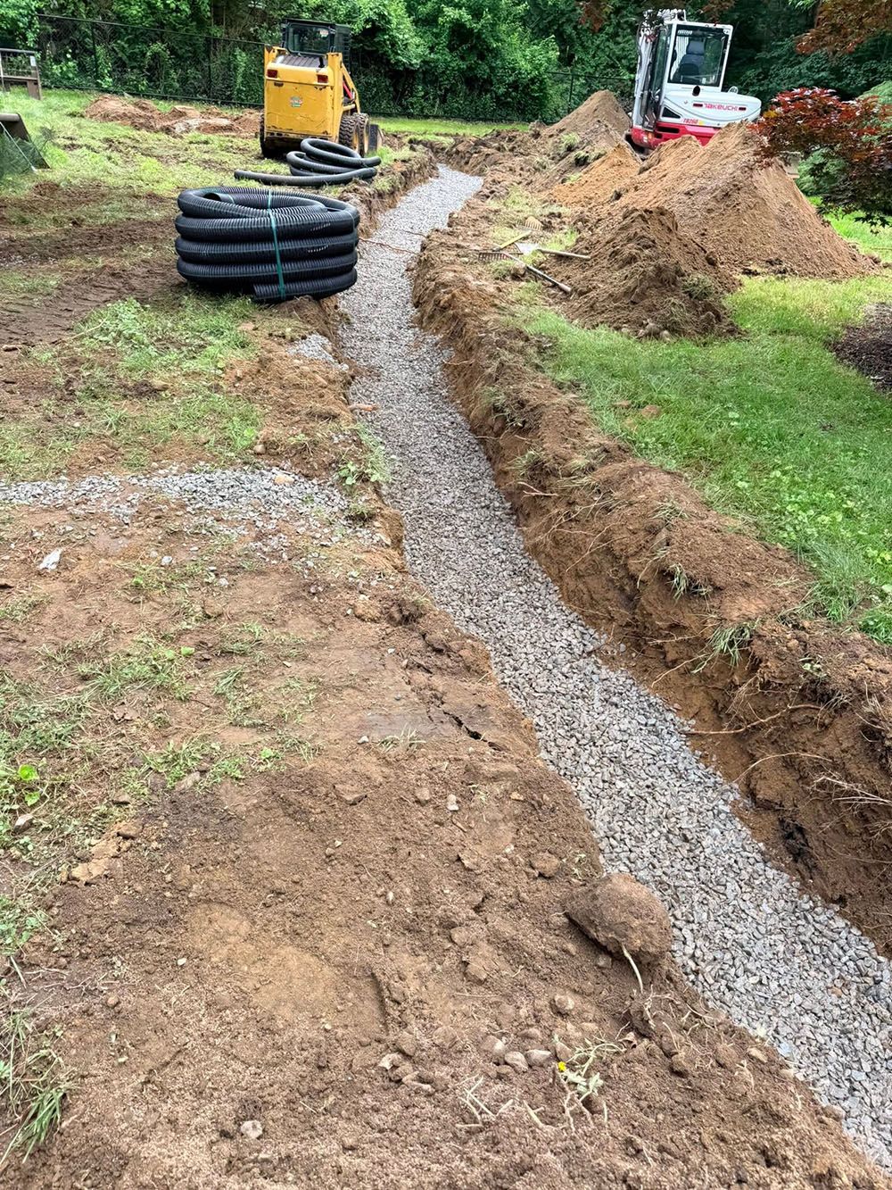 Construction site with gravel trench, stacked black pipe, and a yellow excavator on a dirt slope.