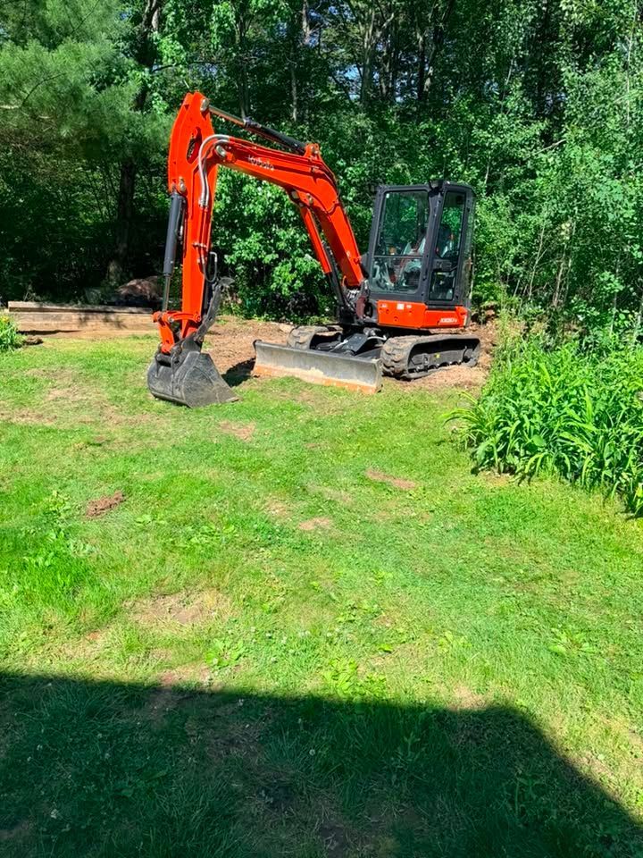 Orange excavator on a grassy yard beside trees, with a bucket lowered in bright sunlight.