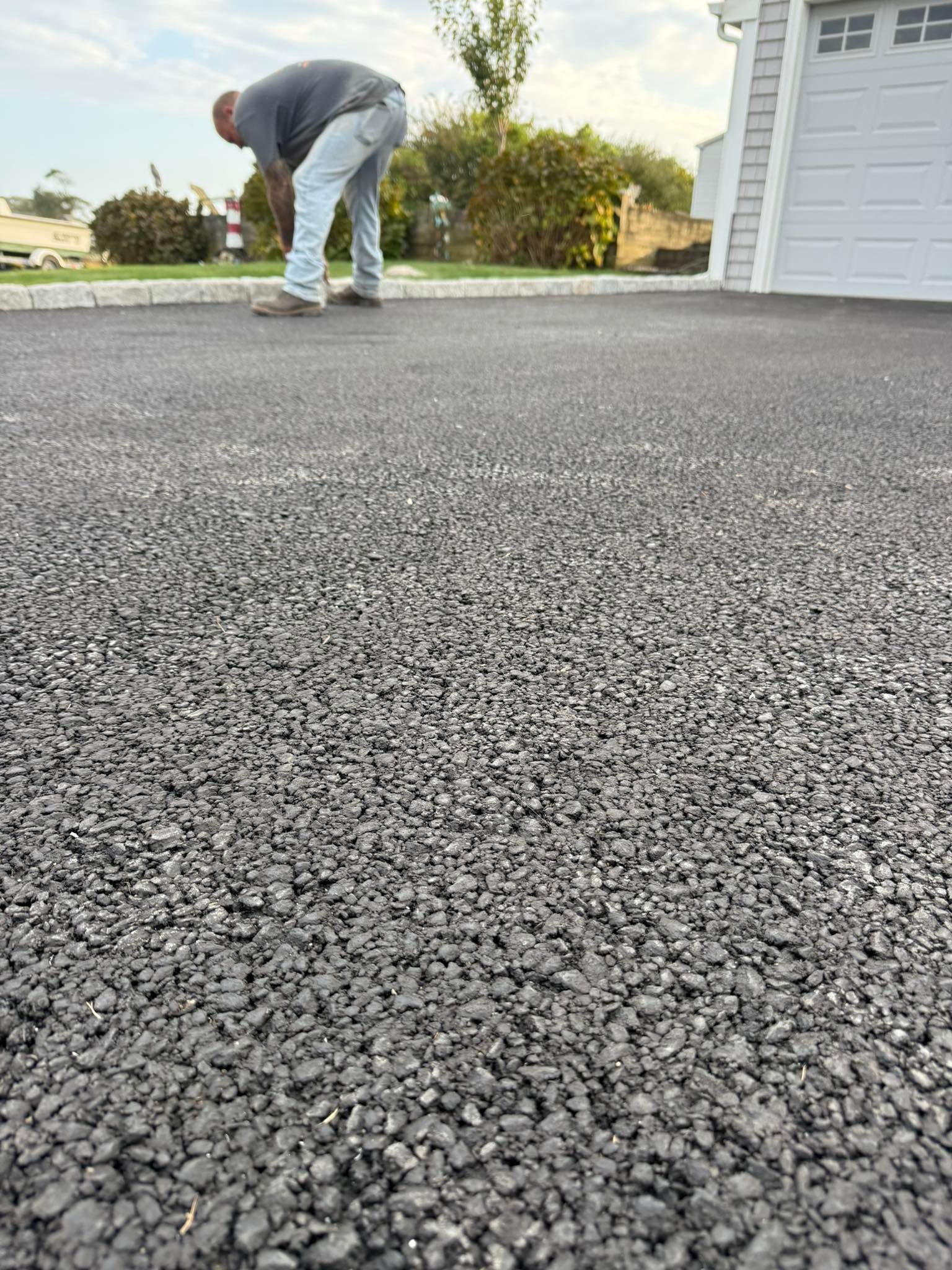 A person bends over to inspect a newly paved, textured asphalt driveway next to a white garage.