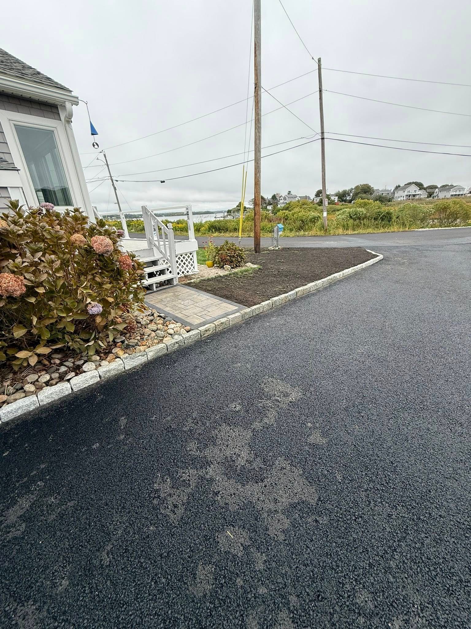 A gravel driveway leads toward a house with a white porch, bordered by stone edging and a garden bed on an overcast day.